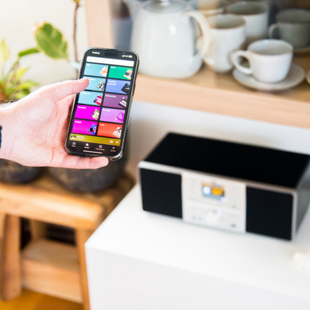 Ręka trzyma telefon z interfejsem aplikacji. System stereo stoi na białej powierzchni. Czajnik i filiżanki na półce.