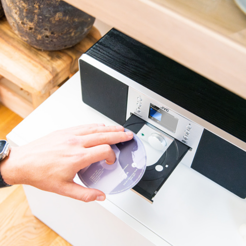 Ręka wkładająca płytę CD do czarno-srebrnego zestawu stereo JVC na białej powierzchni. Drewniany przedmiot w tle.