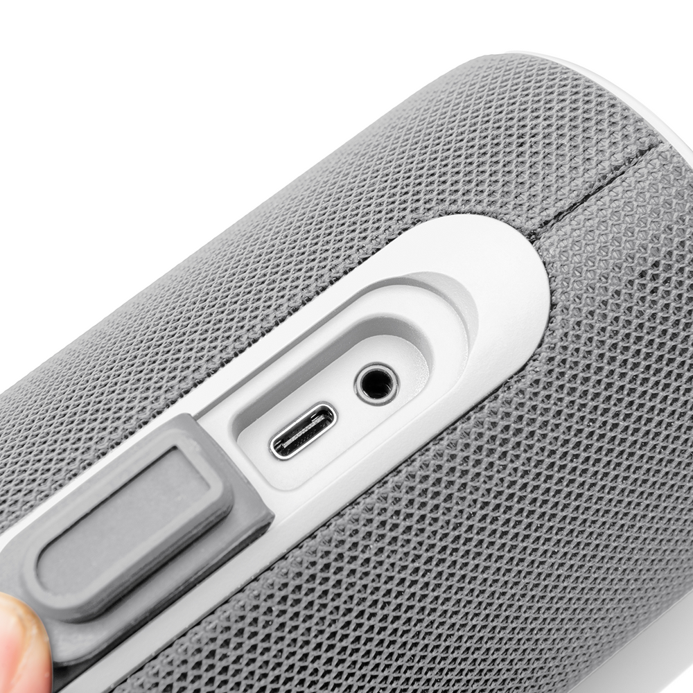 Primo piano di un altoparlante grigio con accenti bianchi. Porte visibili includono USB-C, jack da 3,5 mm e una sezione coperta, tenuta in mano.