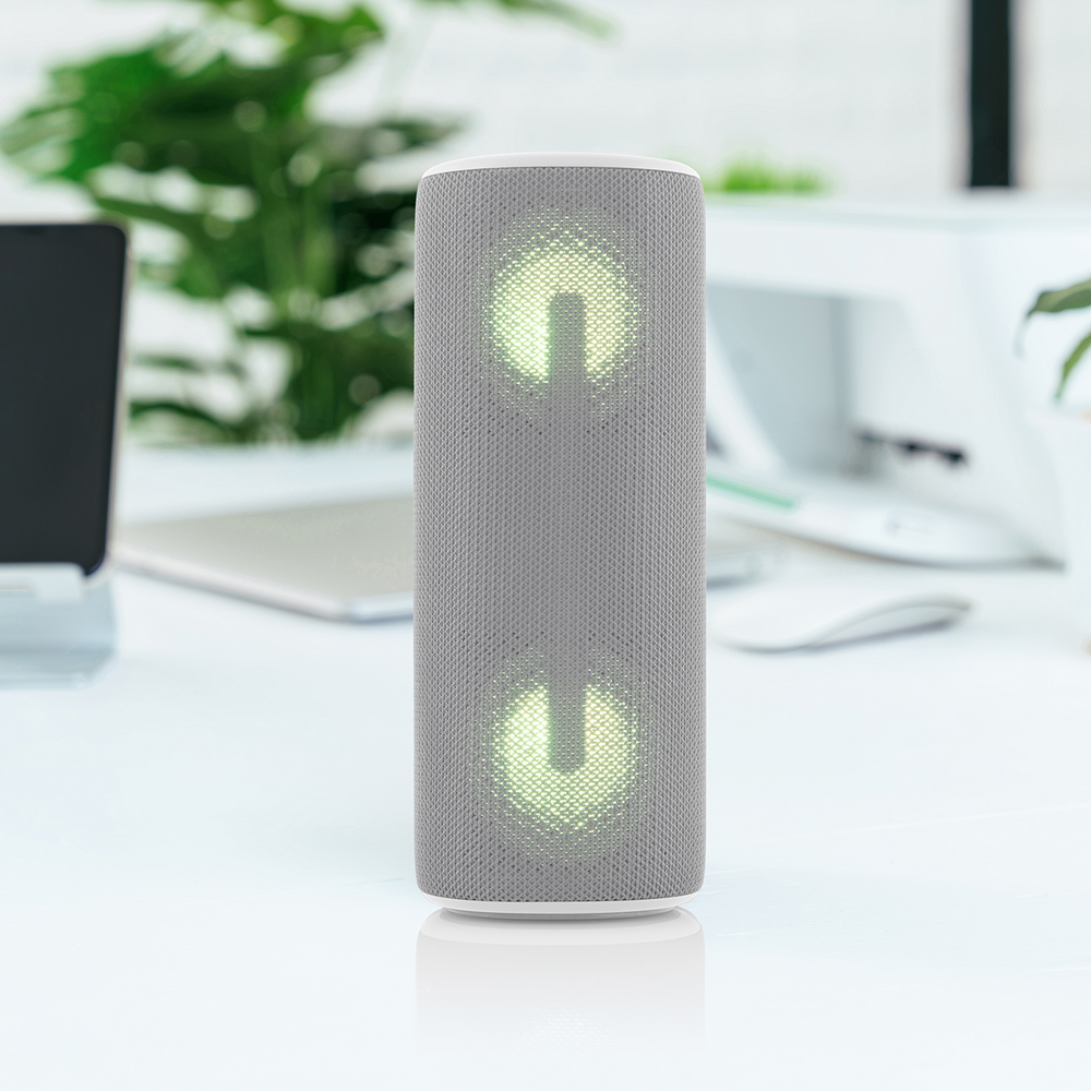 Un altoparlante grigio con simboli di accensione luminosi è su una scrivania bianca. Un laptop, un mouse e delle piante sono sfocati sullo sfondo.