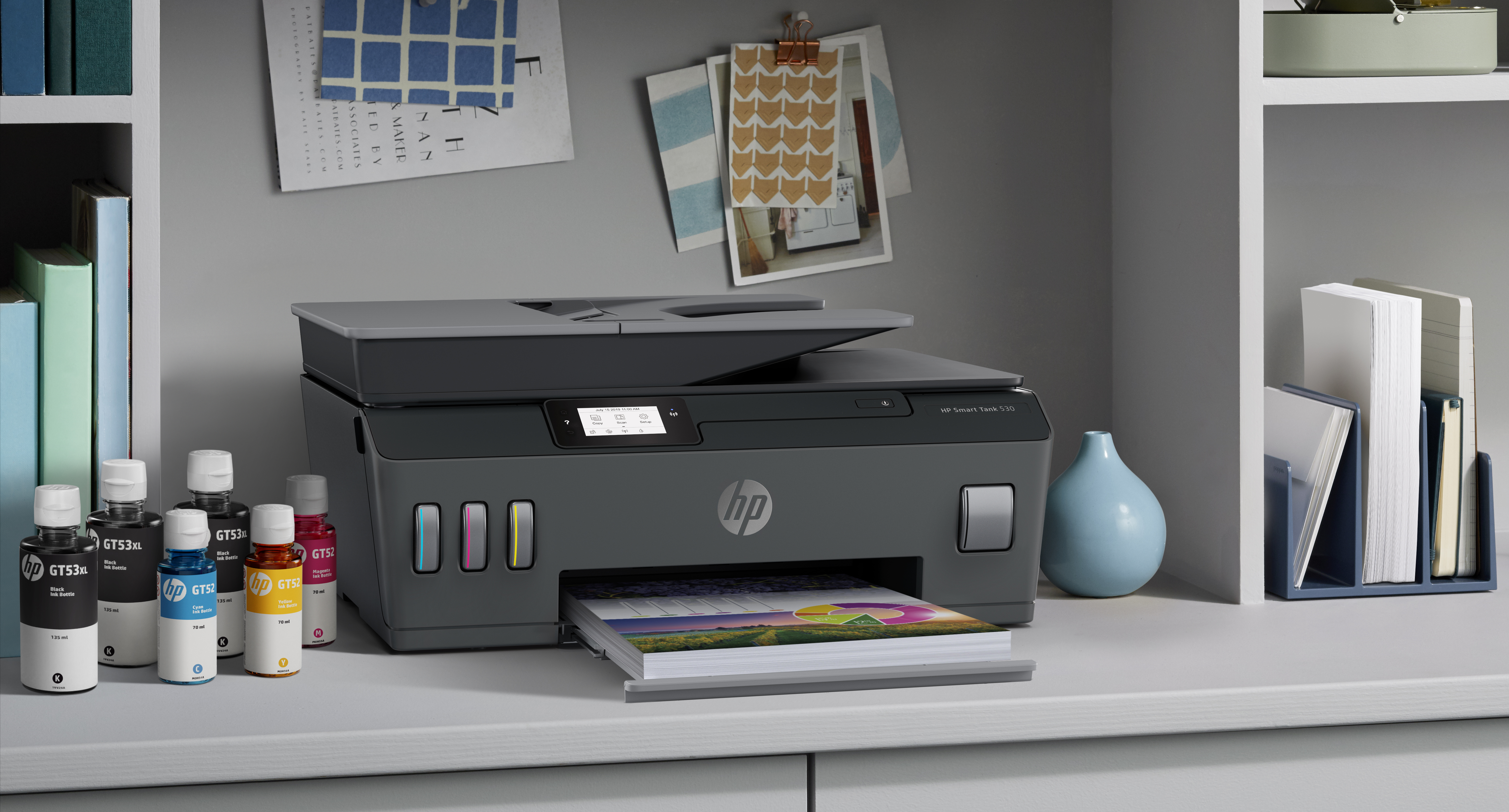 A gray HP Smart Tank 530 printer sits on a desk with ink bottles. There are papers, a blue vase, and art on the wall.