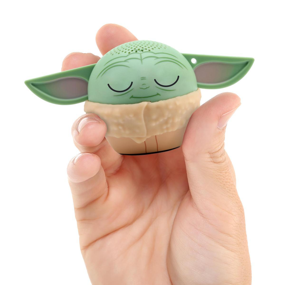 Una mano sostiene un altavoz con forma de Grogu. Cabeza verde, orejas grandes, túnica color beige. El altavoz está sobre un fondo blanco.