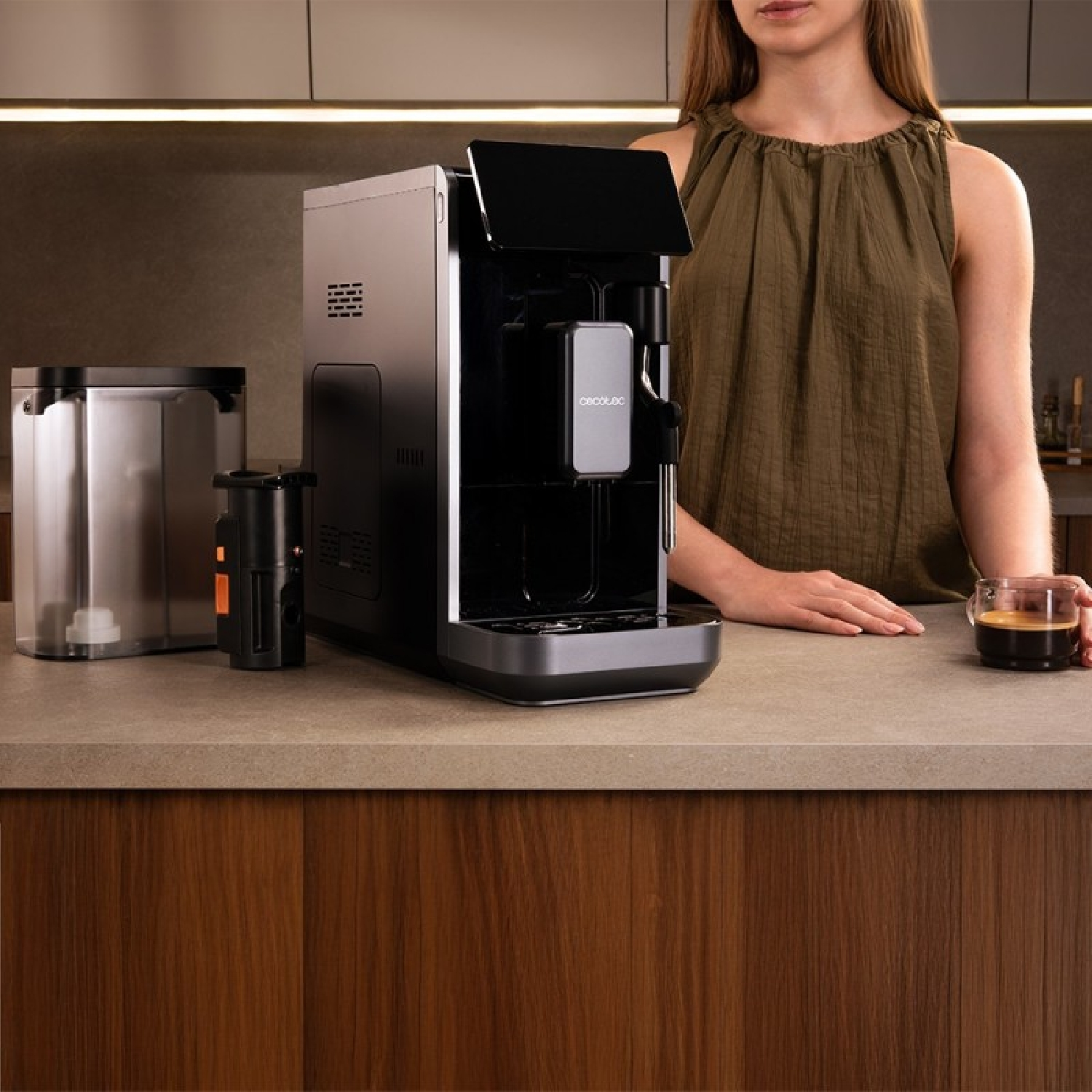 Una donna accanto a una macchina per il caffè, tiene in mano una tazza di caffè. La macchina è su un bancone in cucina.