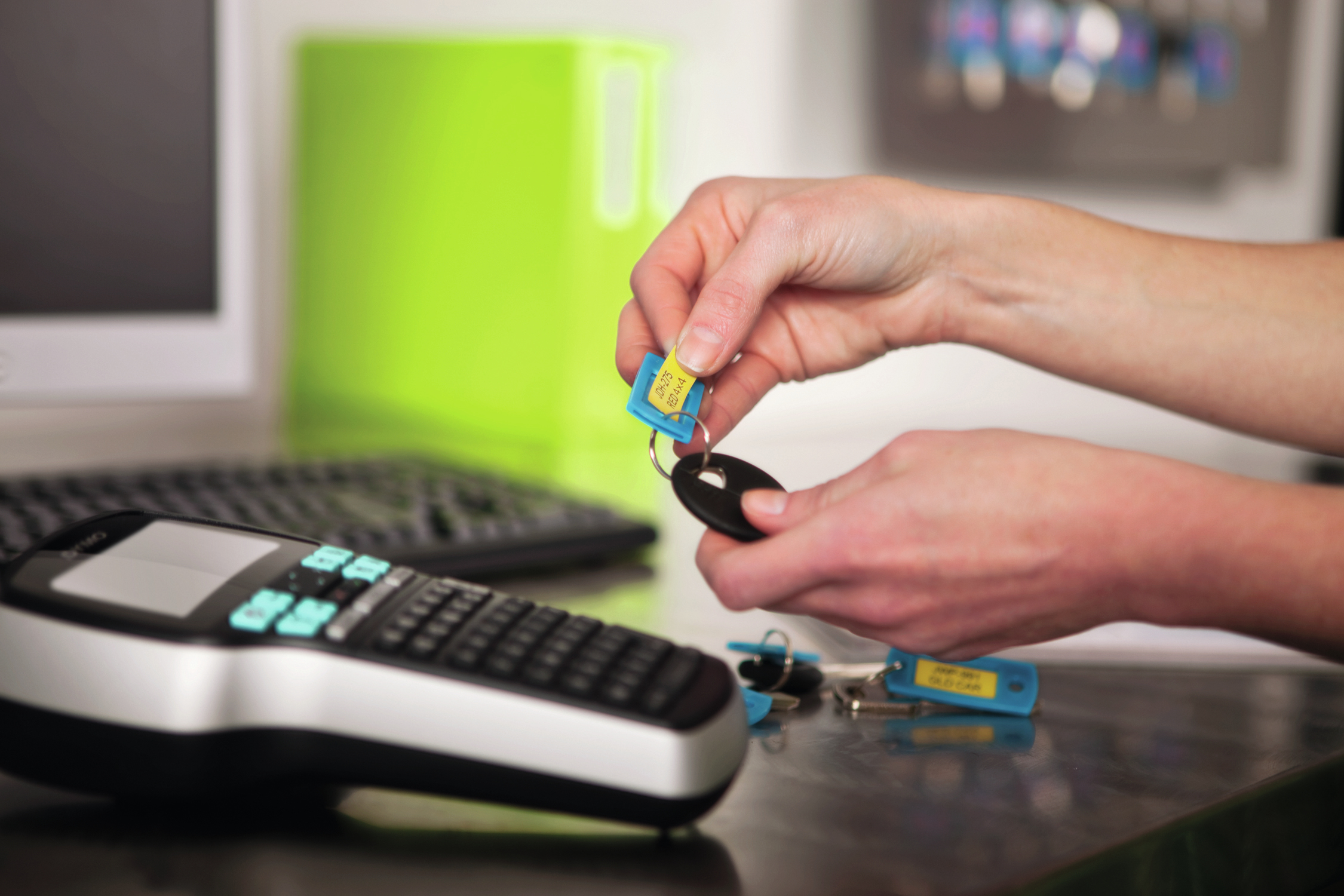 Dłonie trzymają klucze z żółtymi etykietami. Czarny klucz i etykieciarka są na pierwszym planie. Zielony obiekt w tle.
