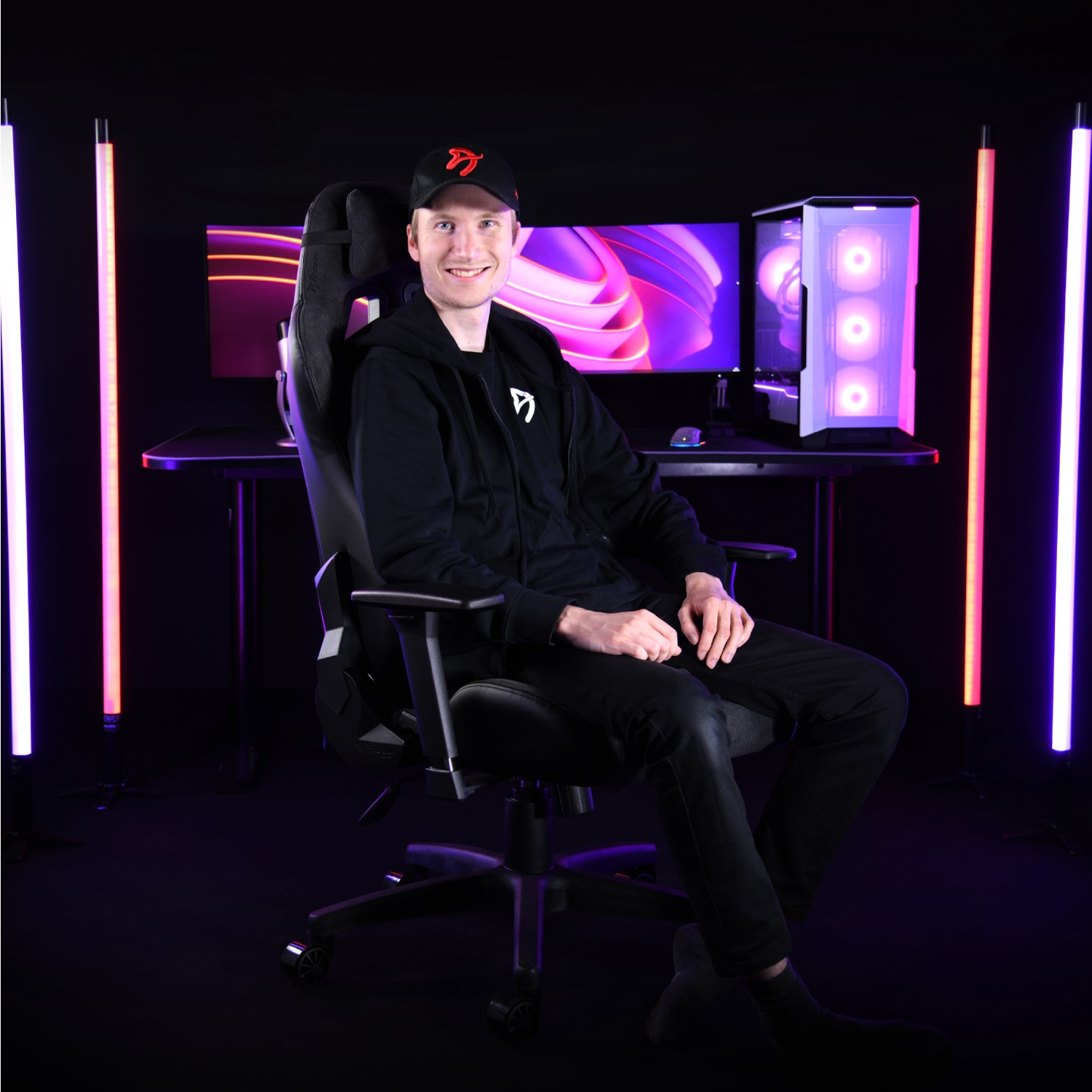Un uomo è seduto su una sedia da gioco, sorridente. Dietro di lui ci sono monitor, un computer e luci colorate. Indossa un cappello e una felpa con cappuccio.