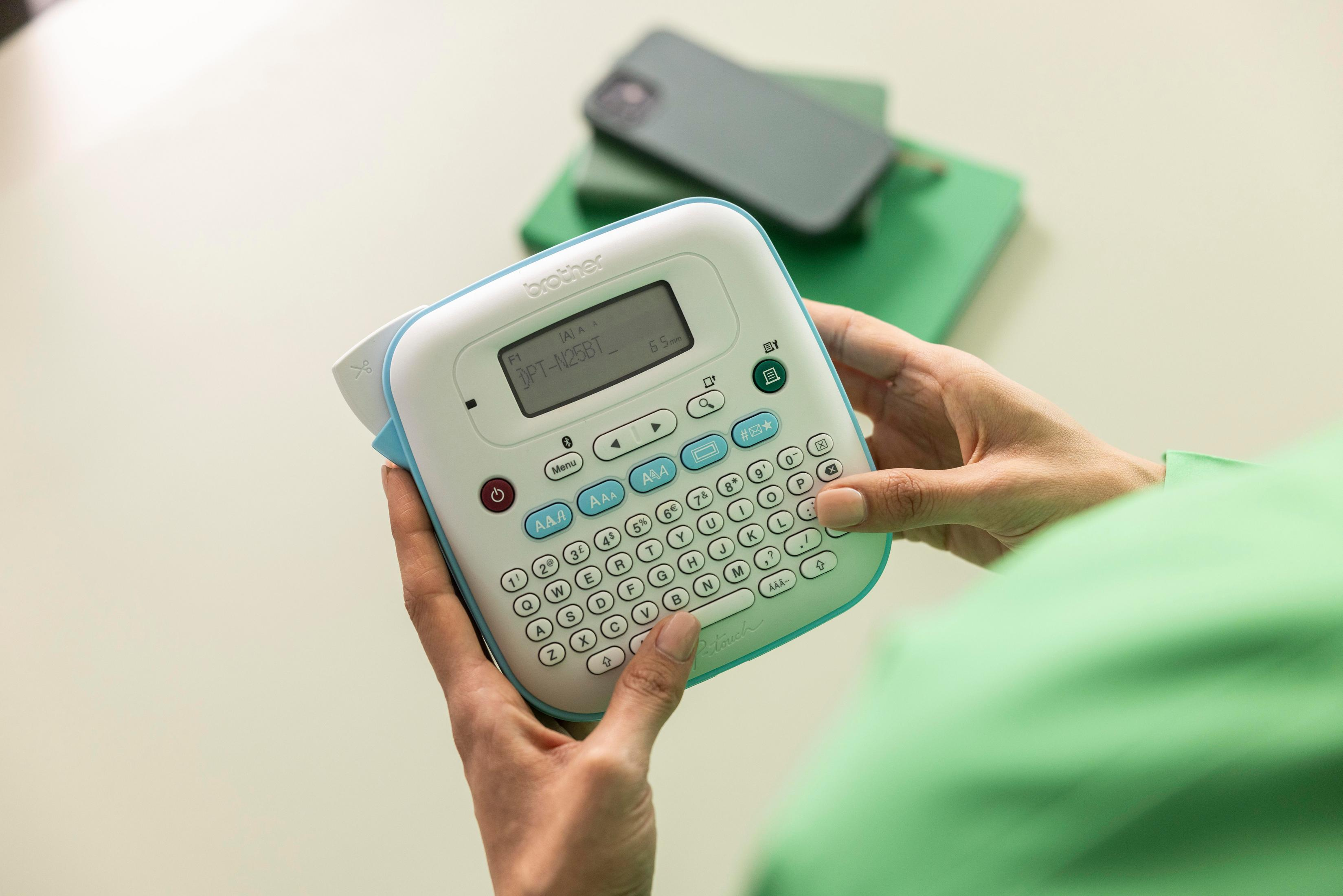 Osoba trzyma etykieciarkę. Telefon i zielony notatnik w tle.