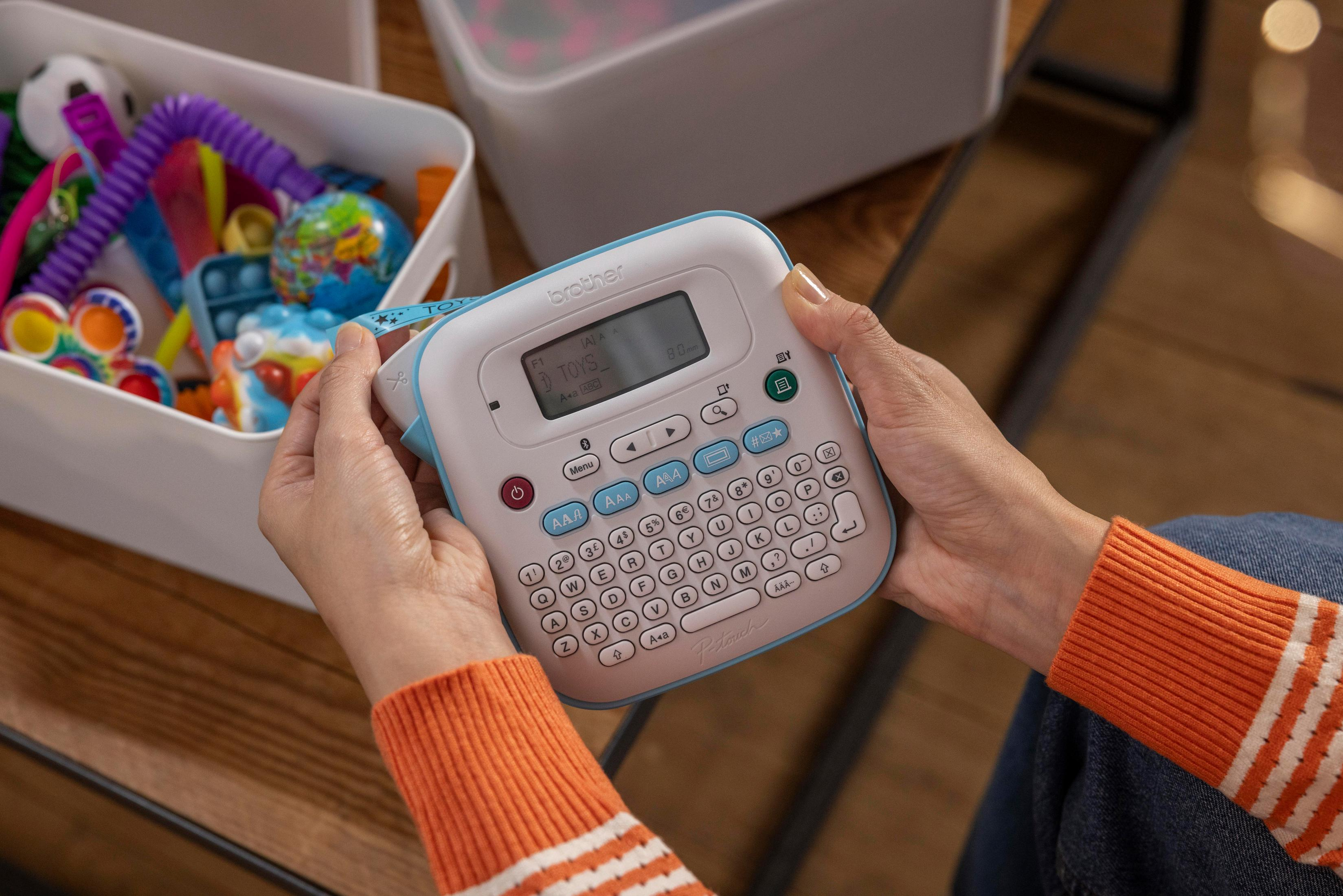 Osoba trzyma etykieciarkę z napisem 'TOYS'. Zabawki w białym pojemniku w tle. Osoba ma na sobie pomarańczowy sweter.