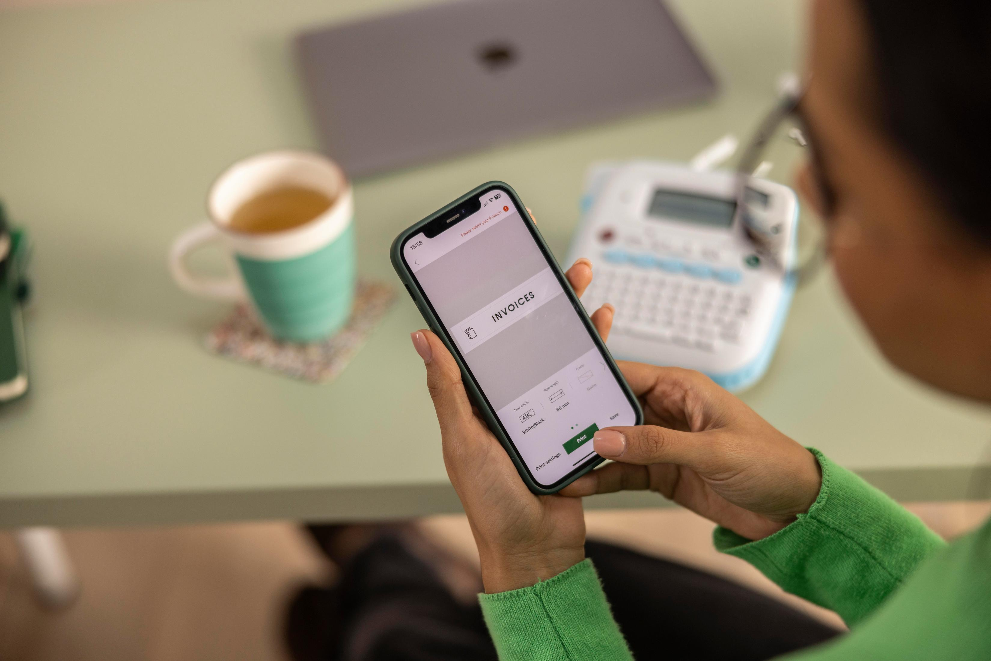 Osoba trzyma telefon z aplikacją do fakturowania, w pobliżu drukarki etykiet i filiżanki herbaty.