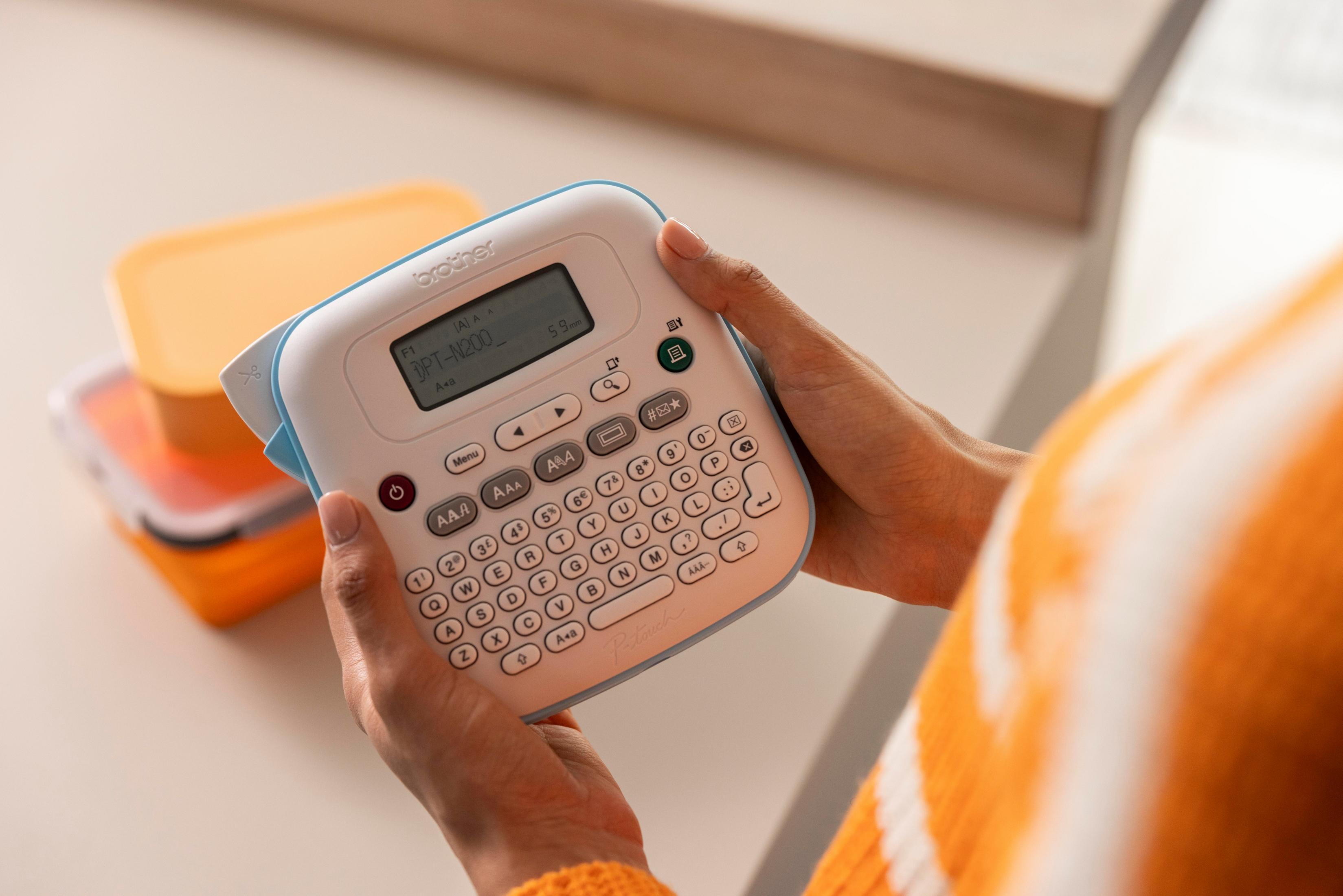 Osoba trzyma etykieciarkę Brother. W tle znajdują się pomarańczowe pojemniki. Osoba ma na sobie pomarańczową górę.