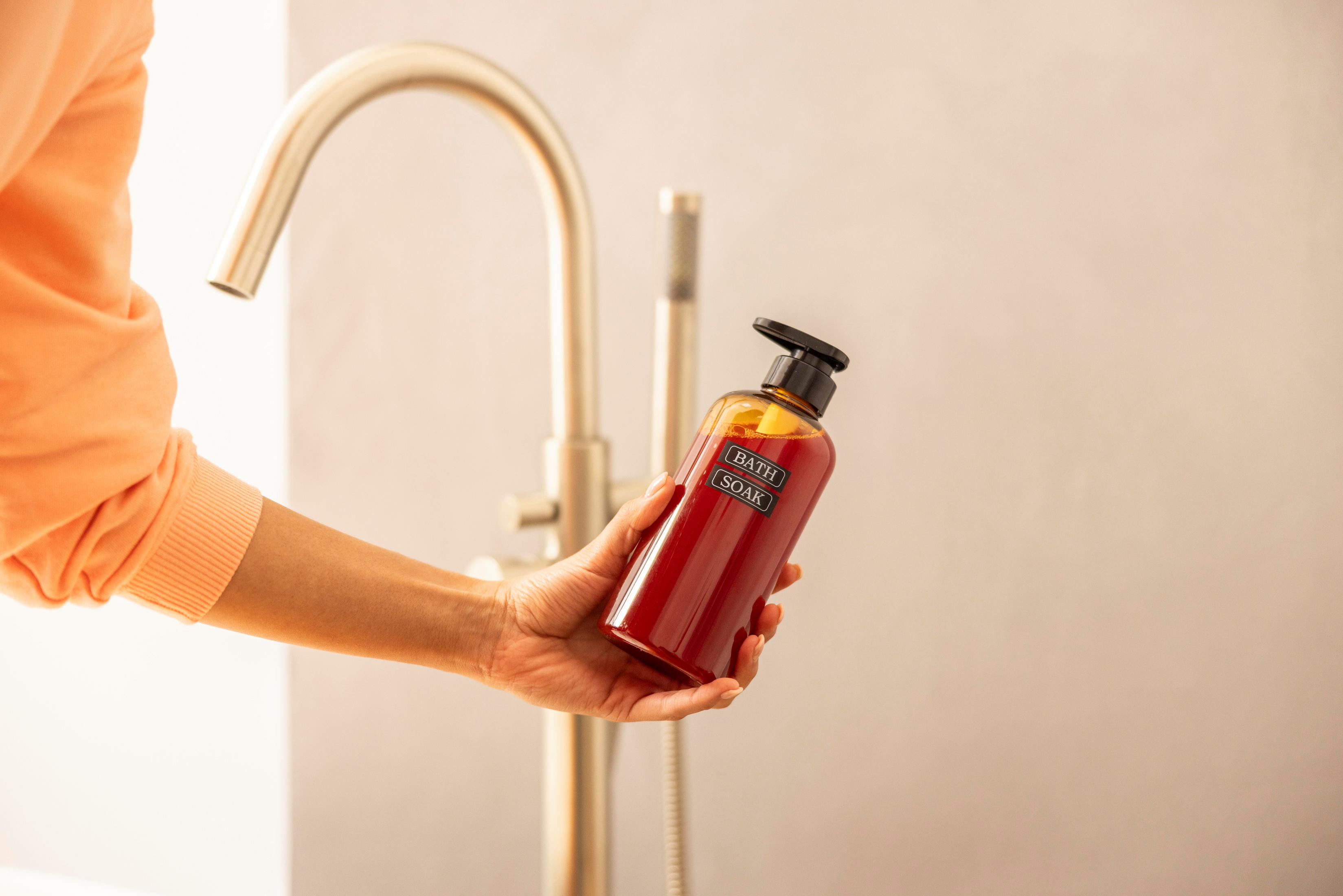 Ręka trzyma butelkę do kąpieli w pobliżu kranu. Butelka jest czerwona z czarną zakrętką. Osoba ma na sobie pomarańczowy.