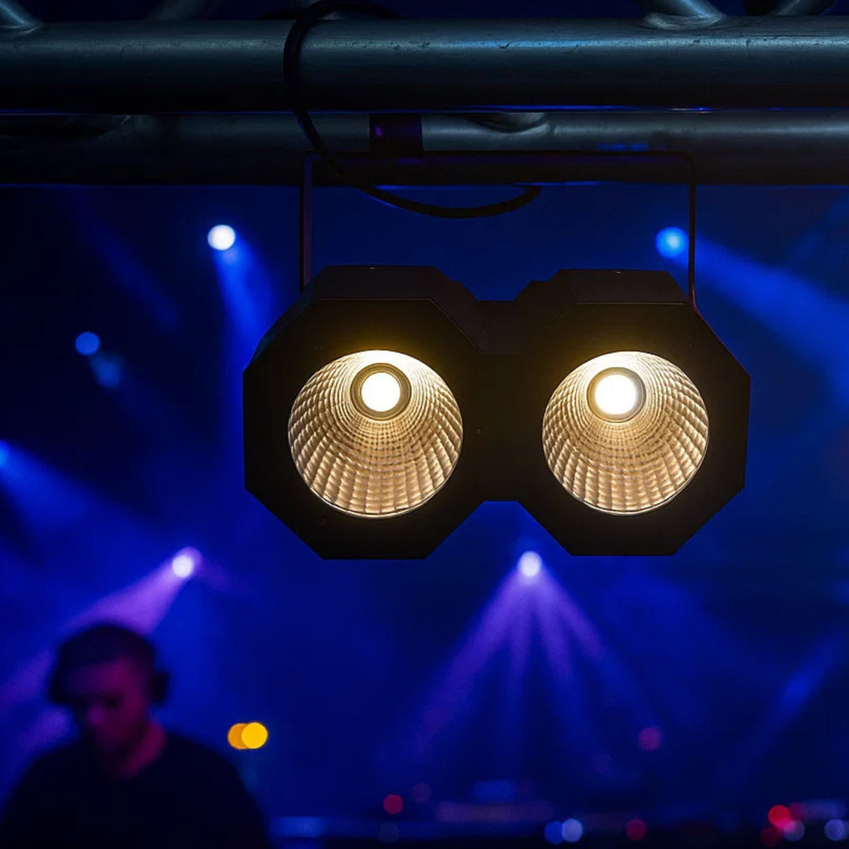 Zwei Bühnenlichter mit hellen Glühbirnen hängen, im Hintergrund ein DJ. Bläuliche und violette Lichter füllen den Hintergrund.