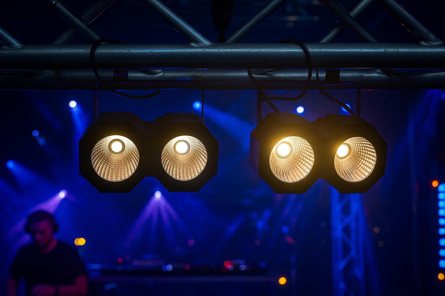 Bühnenbeleuchtung hängt an Metallträgern. Zwei Sätze Doppellichter leuchten mit hellem gelbem Licht. Ein DJ ist im Hintergrund verschwommen.