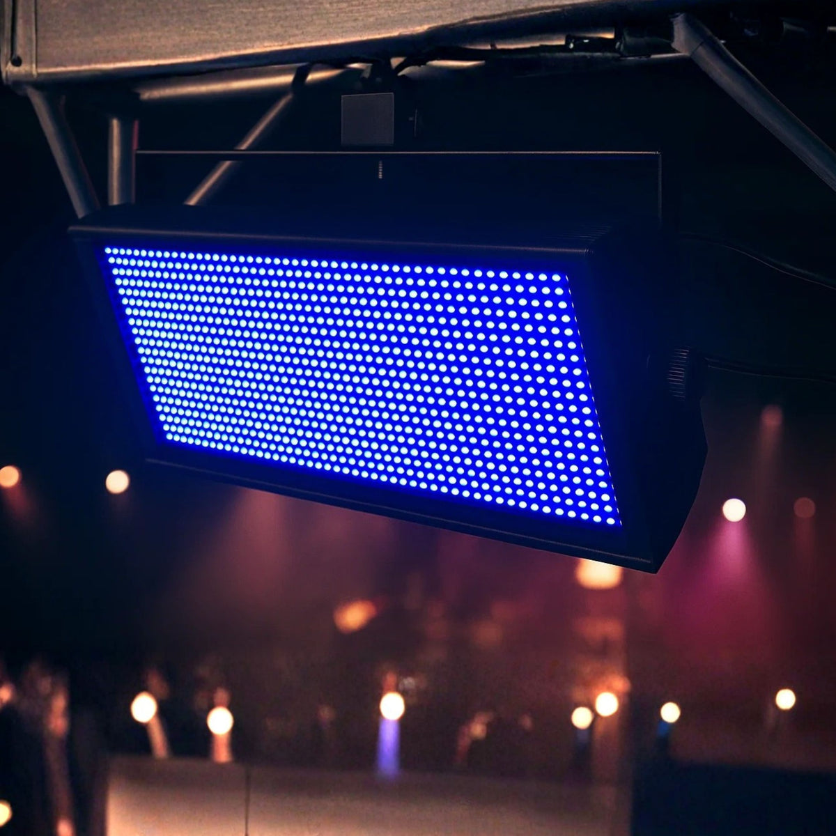 Ein rechteckiges LED-Panel mit blauen Lichtern hängt an Metallträgern. Der Hintergrund zeigt verschwommene Bühnenlichter und Silhouetten.