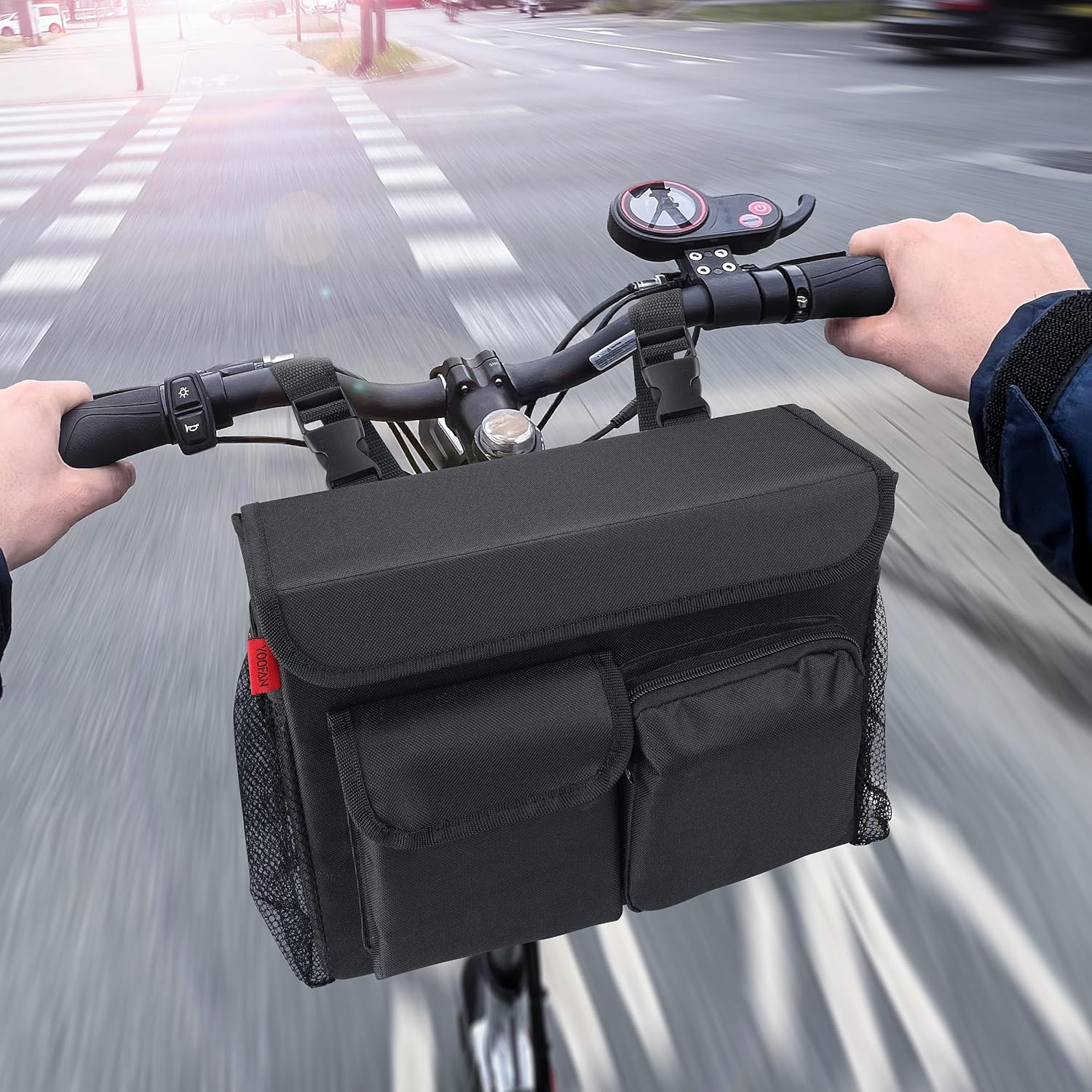 Eine Person fährt mit einem Roller auf einer Straße. Eine schwarze Tasche ist am Lenker befestigt. Die Straße hat einen Zebrastreifen.