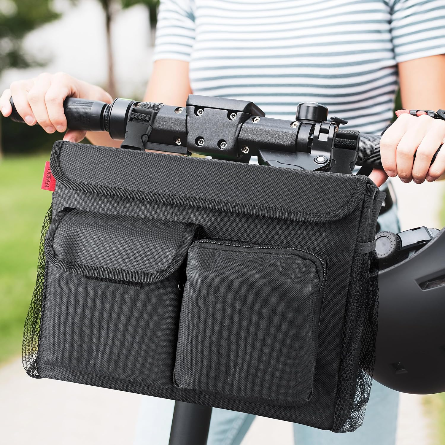 Eine Person hält einen Rollerlenker mit einer schwarzen Tasche. Die Tasche hat Taschen. Ein Helm ist an der Seite. Hintergrund ist grün.