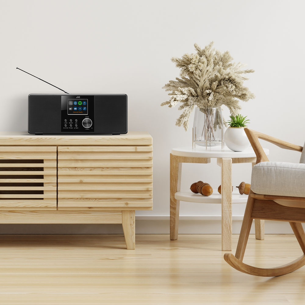 Czarny radioodtwarzacz JVC stoi na drewnianej szafce. Wazon z suszonymi kwiatami i roślina na stoliku. Obecny jest fotel bujany.