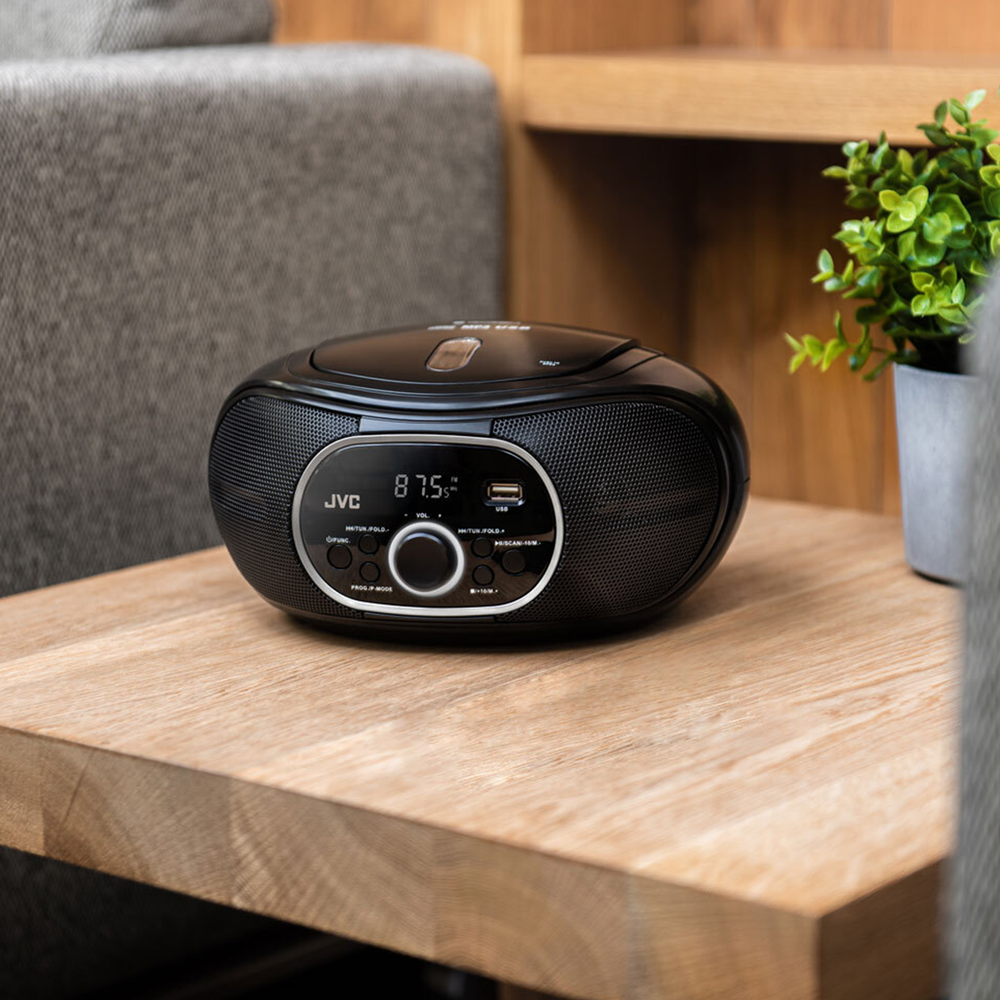 Czarny boombox JVC stoi na drewnianym stole. Po prawej stronie mała roślina w szarej doniczce. Szara kanapa w tle.