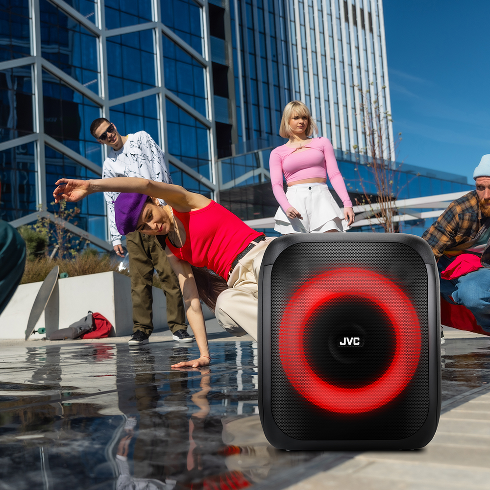 Kobieta tańczy w pobliżu głośnika JVC z czerwonym światłem. Ludzie stoją w pobliżu. W tle nowoczesny budynek.