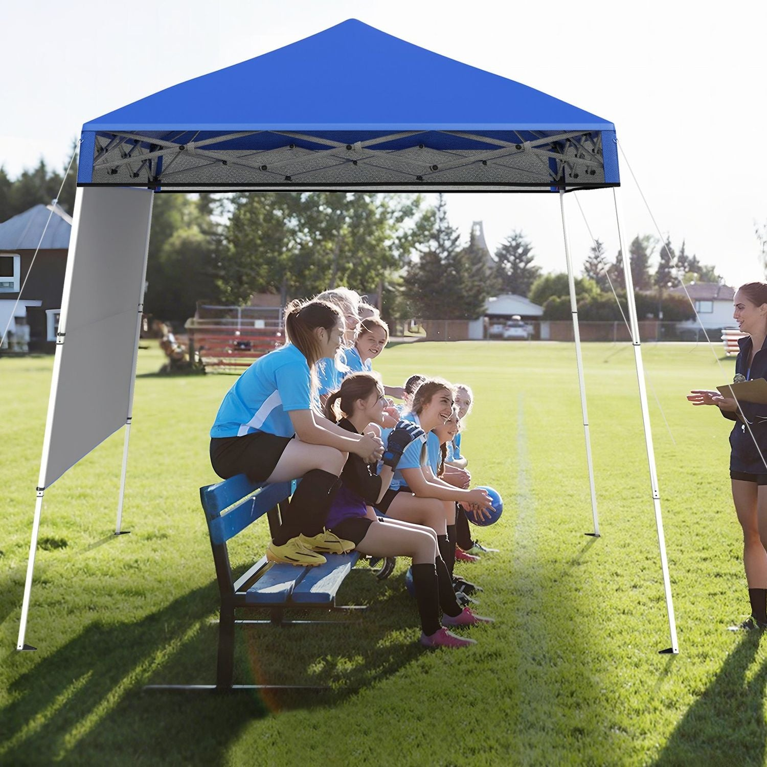 Equipo de fútbol bajo un toldo azul. Jugadores sentados en un banco, entrenador con portapapeles. Campo verde, casas al fondo.