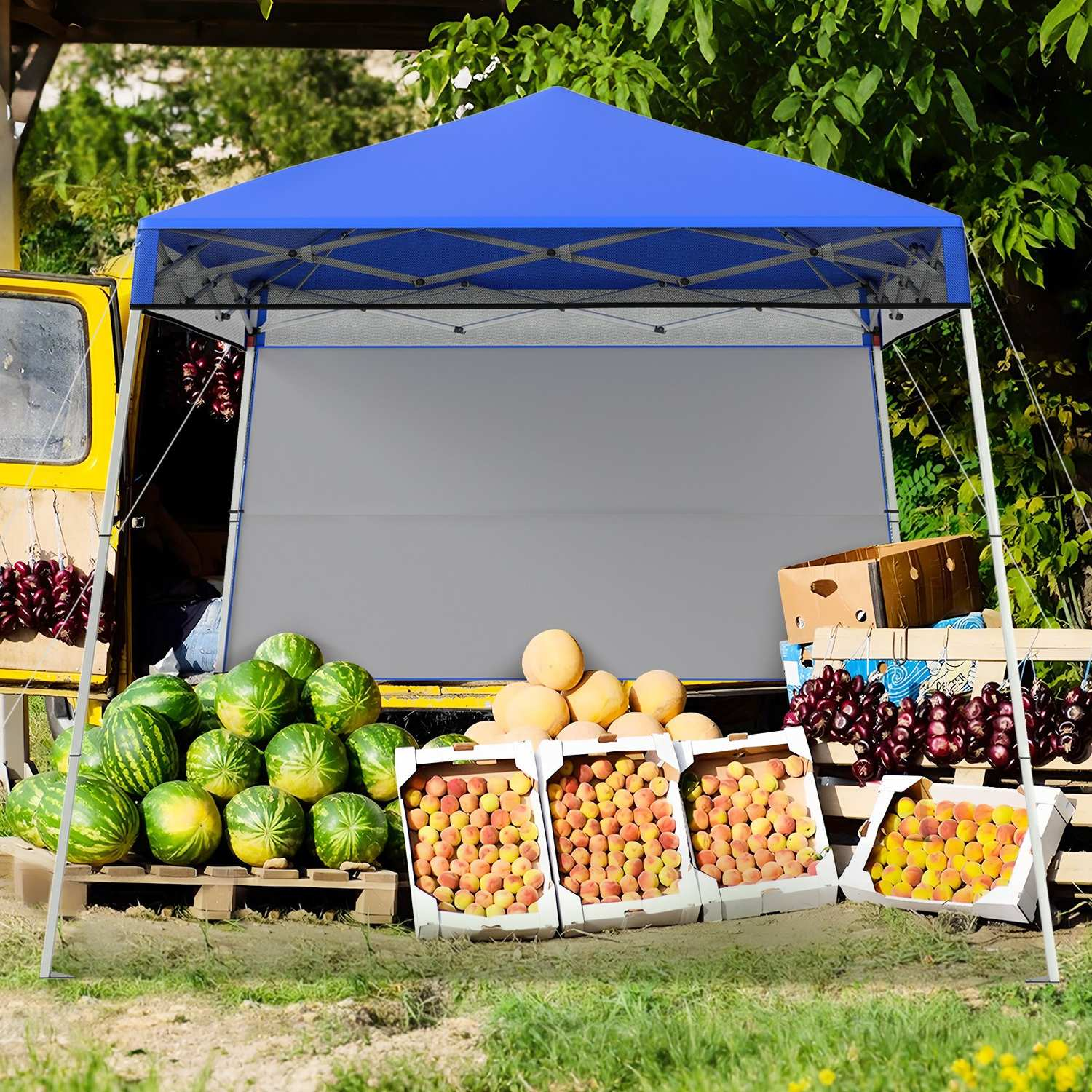 Un puesto de frutas con toldo azul exhibe sandías, melocotones y otras frutas. Un vehículo amarillo está al fondo.