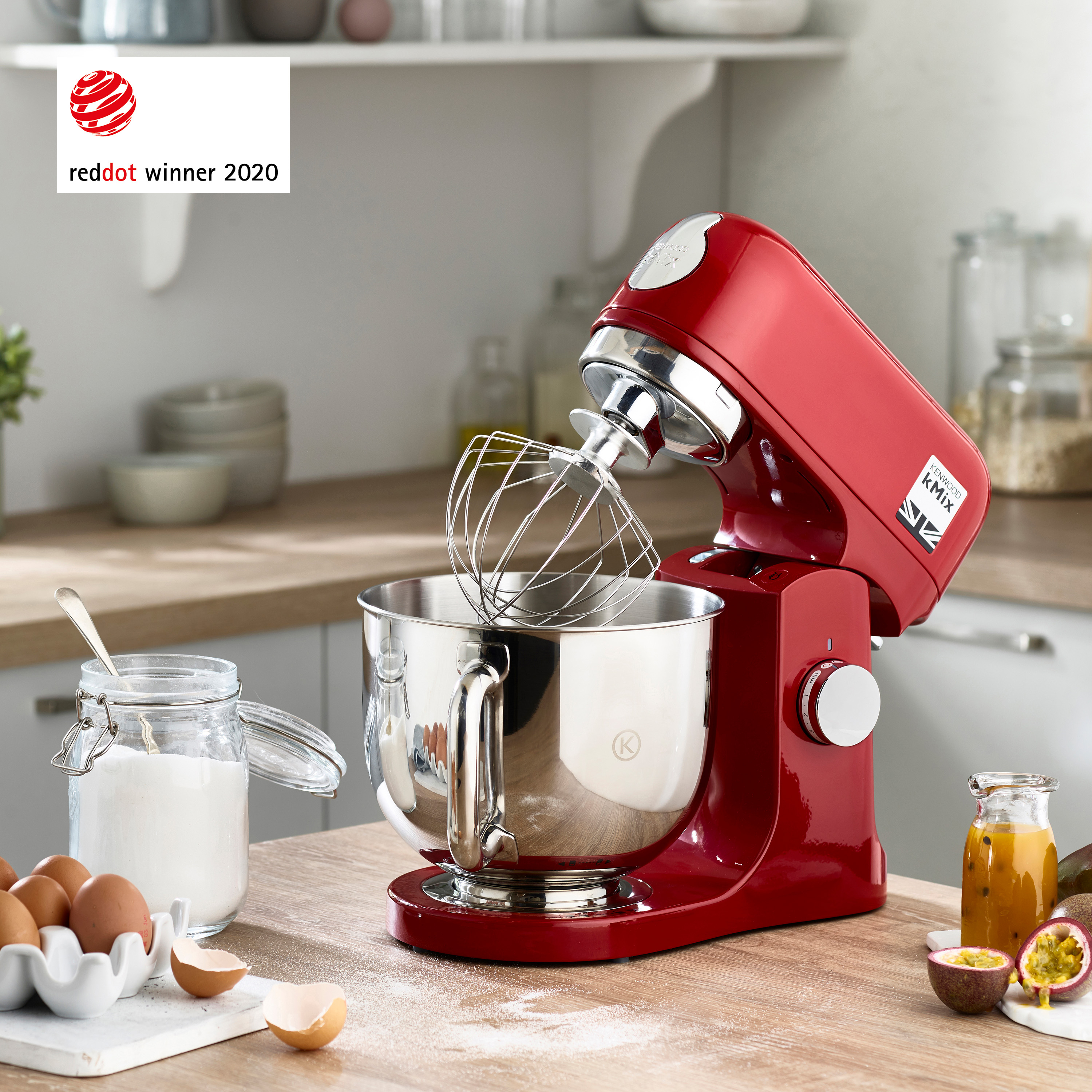 Ahşap bir masada yumurta ve şeker gibi malzemelerle birlikte kırmızı bir Kenwood Kmix stand mikser. Red Dot kazananı 2020.