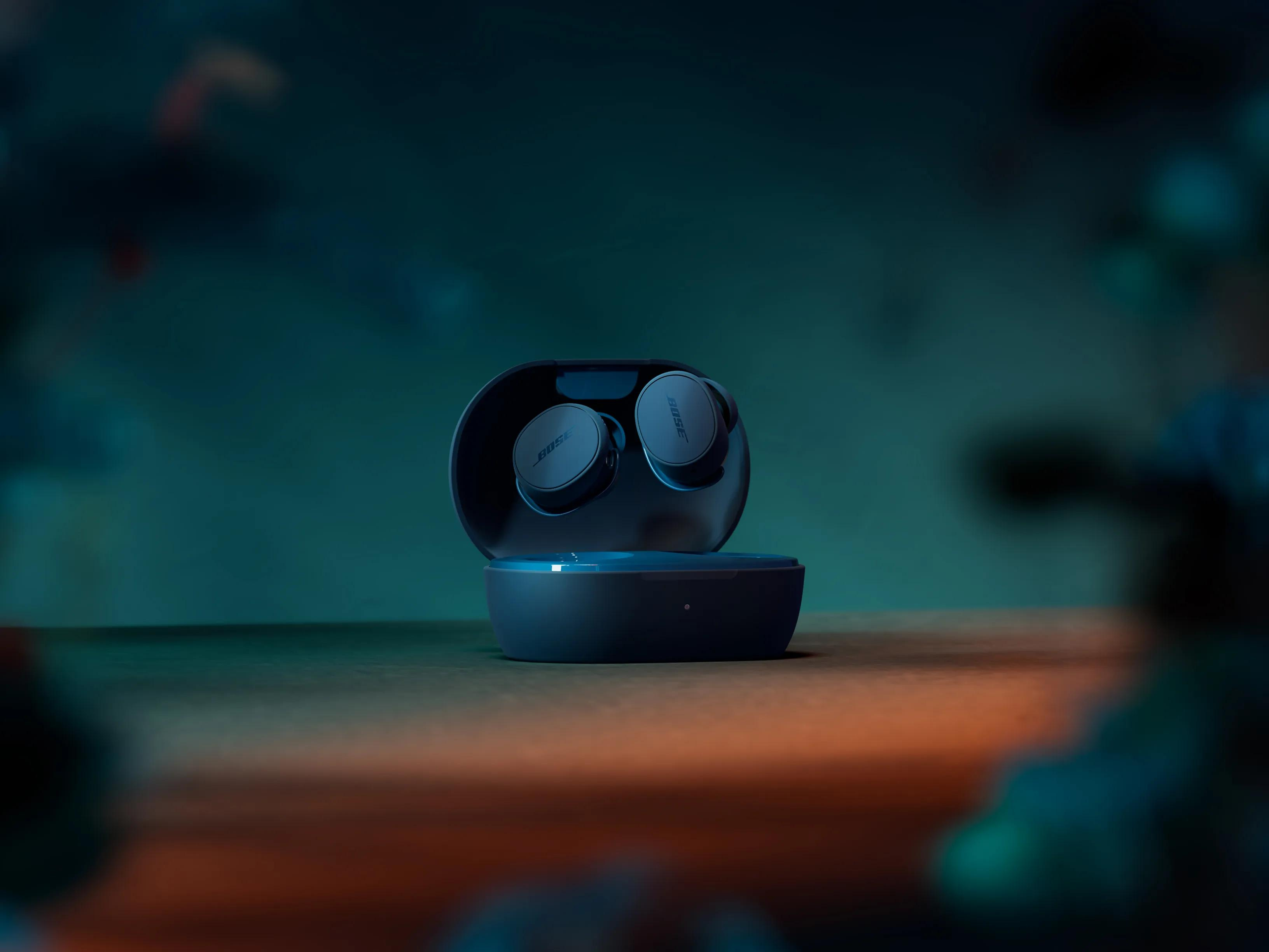 Auriculares Bose en un estuche de carga abierto. Auriculares y estuche azul oscuro sobre una superficie marrón con fondo verde azulado.