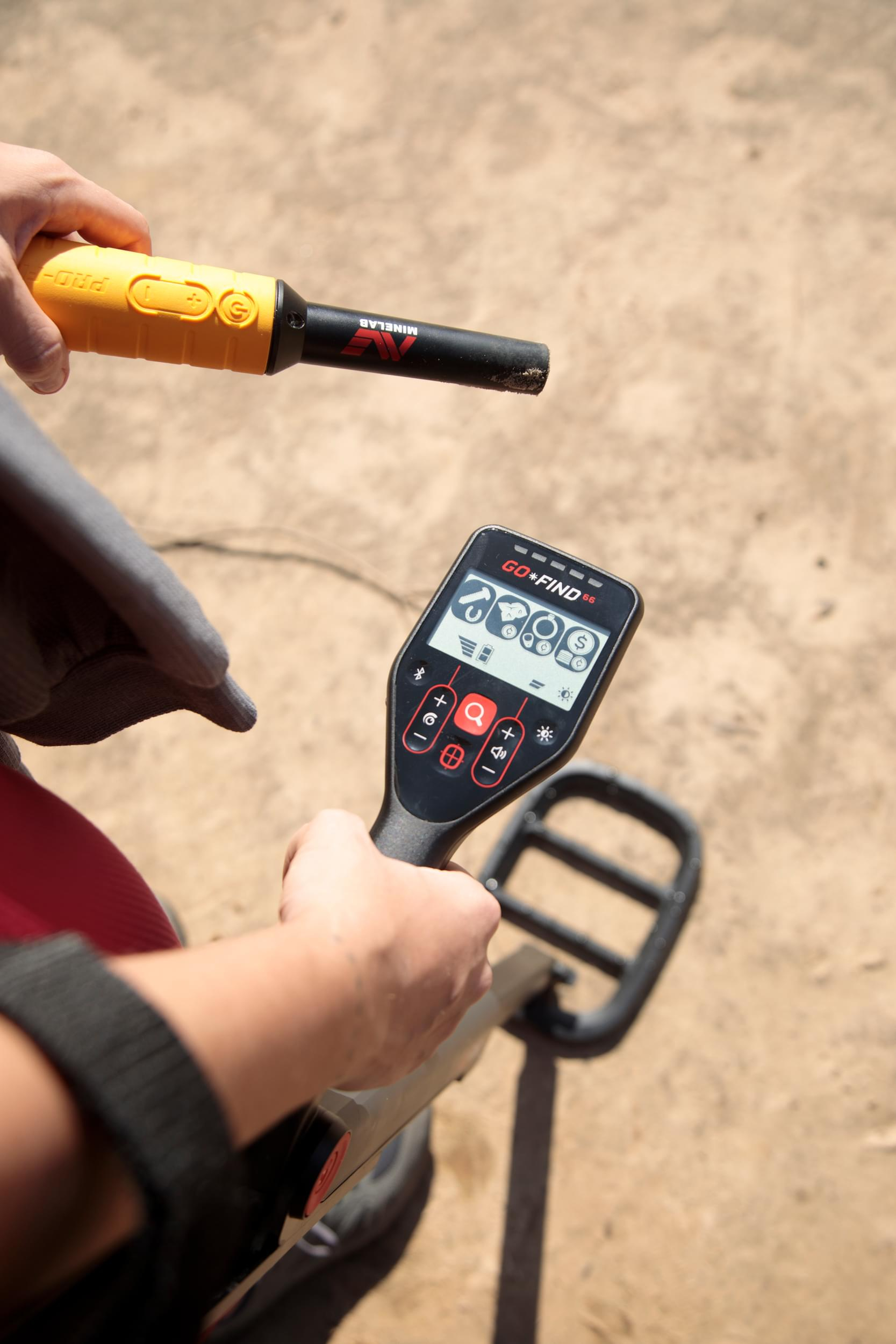 Una persona tiene in mano un metal detector, mostrando il display e la sonda. Lo sfondo è sabbia sfocata.