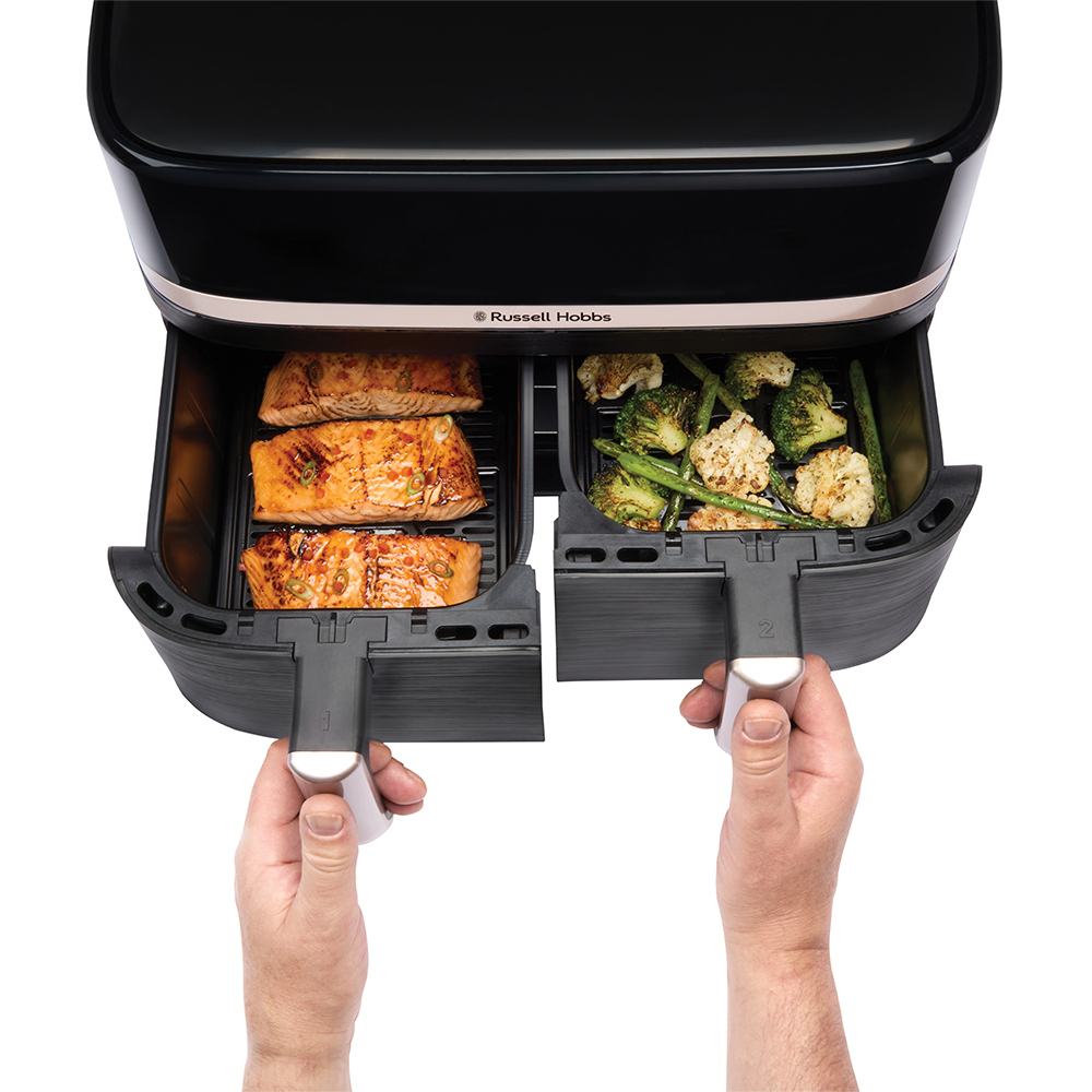 Frytownica powietrzna z łososiem i warzywami. Dwie ręce wyciągają tace. Frytownica jest czarna.