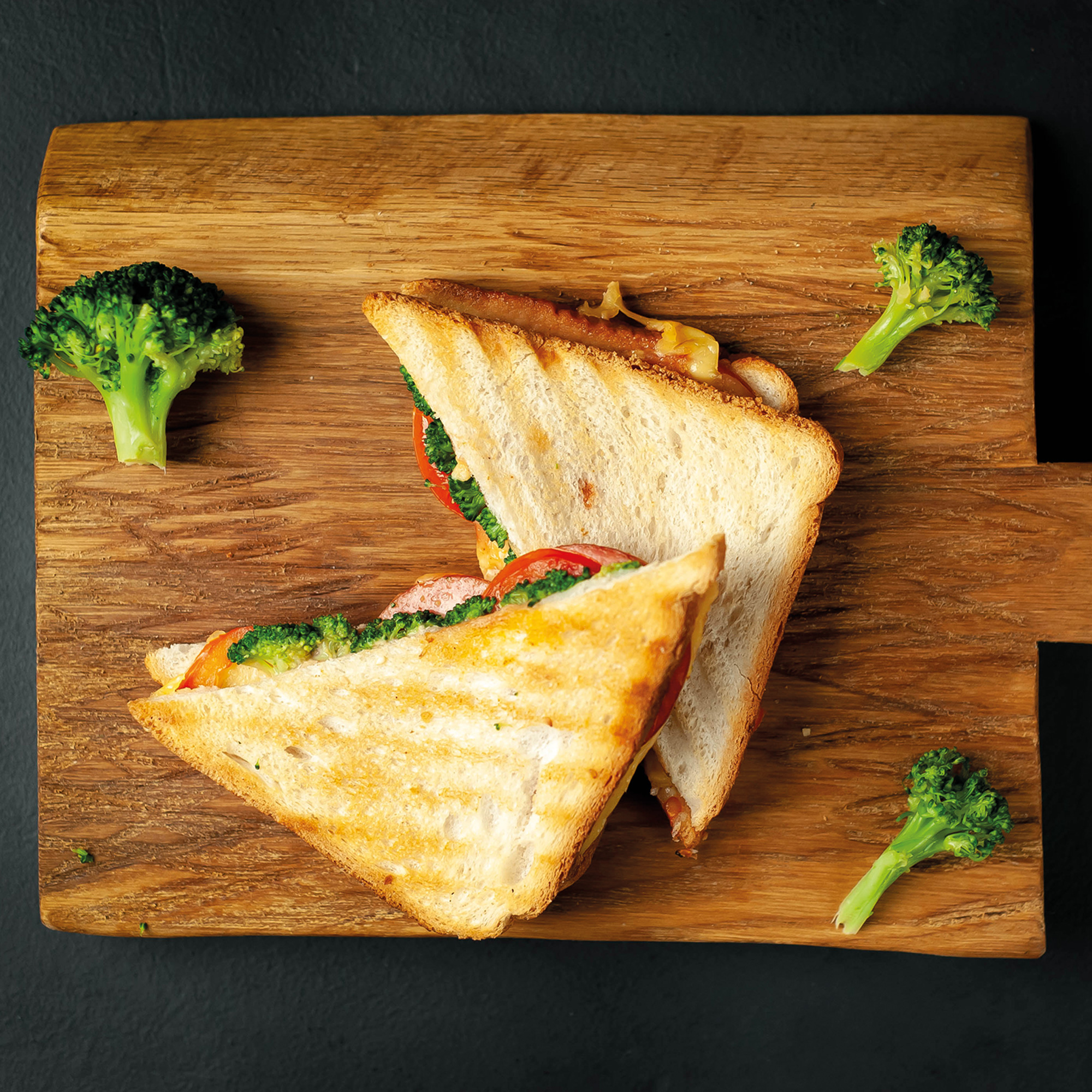 Ein getoastetes Sandwich mit Brokkoli und Tomaten auf einem Holzbrett. Brokkoliröschen umgeben das Sandwich auf schwarzem Grund.
