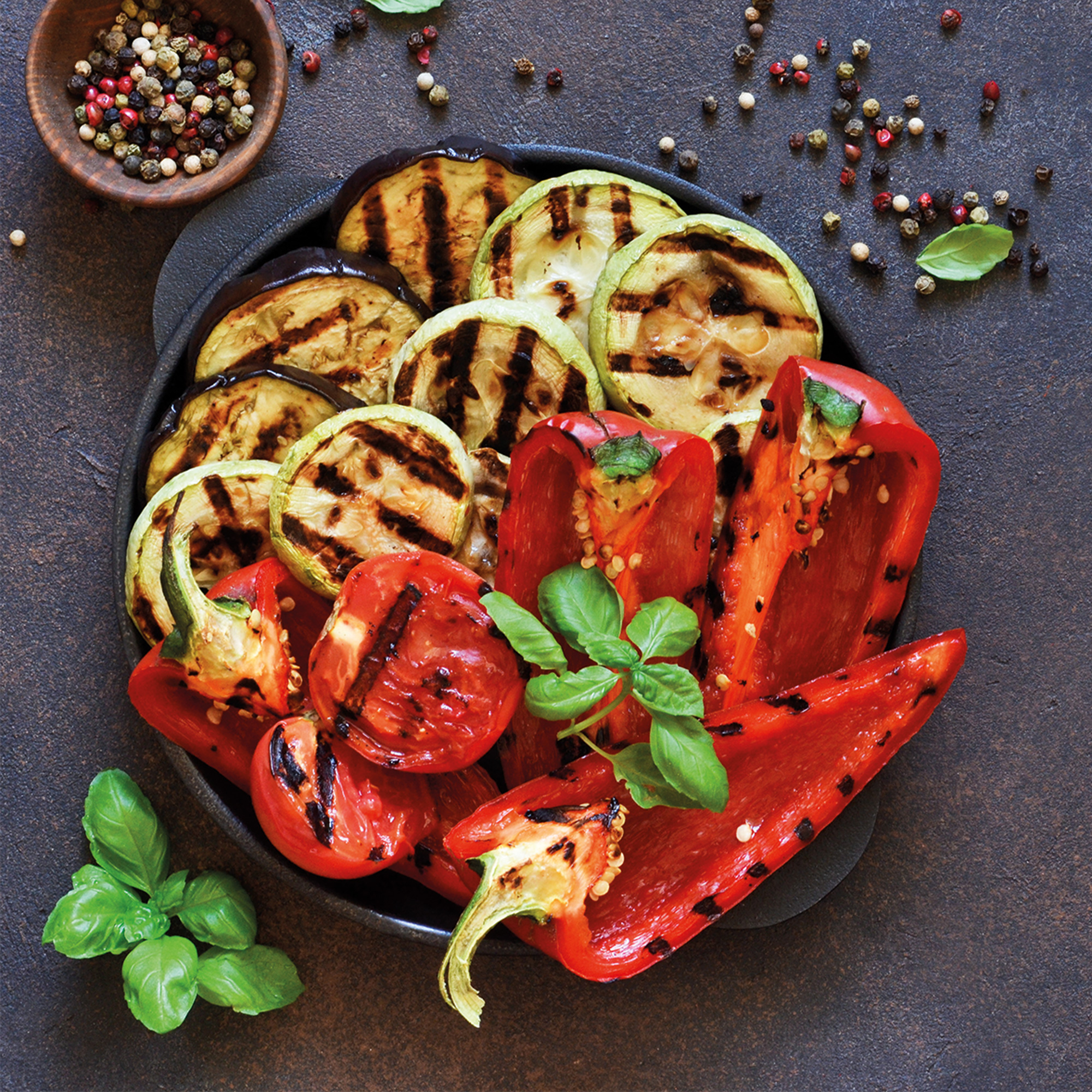 Gegrilltes Gemüse: Zucchini, Aubergine, Paprika und Tomaten in einer schwarzen Pfanne mit Basilikumblättern und Pfefferkörnern.
