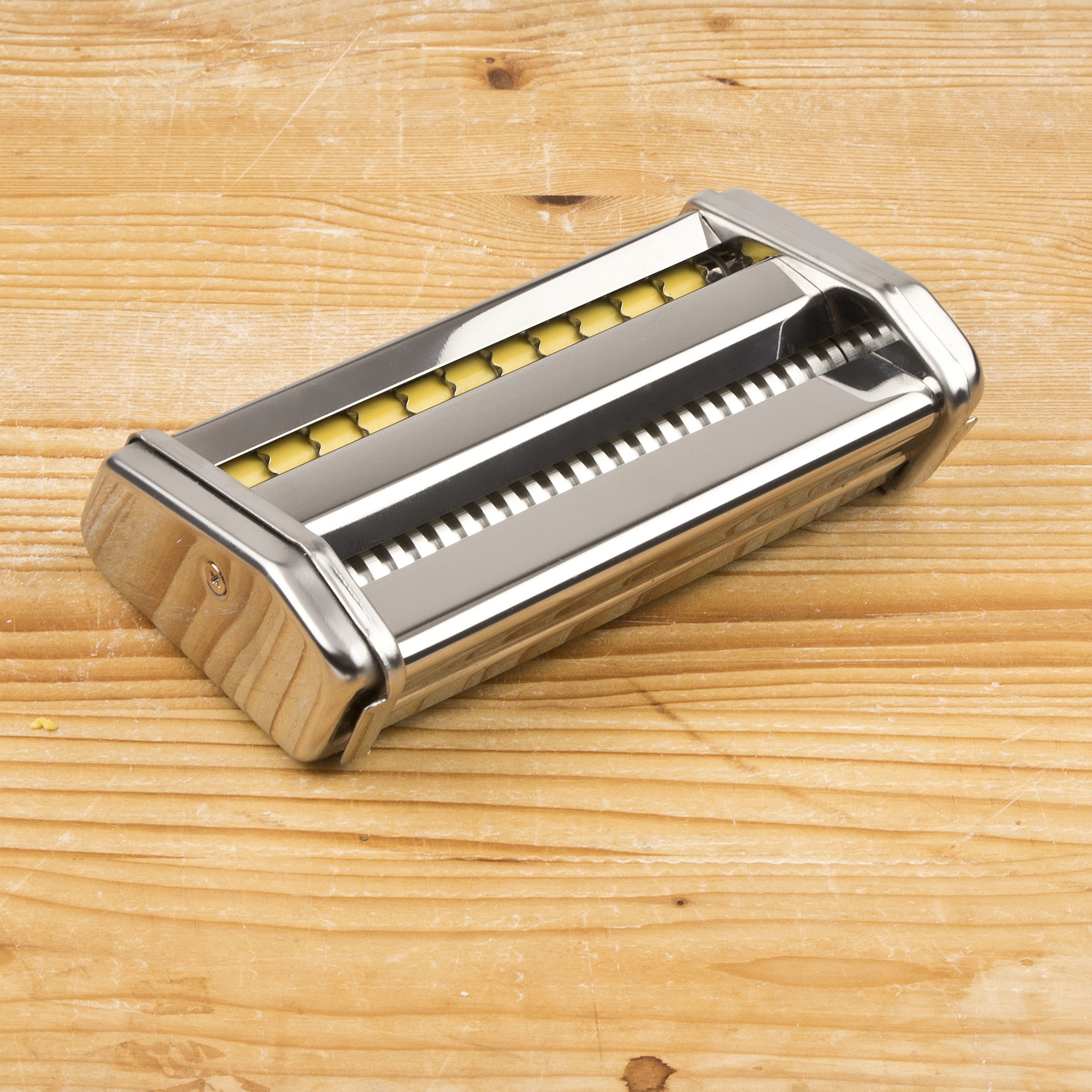 Eine Metall-Nudelmaschine liegt auf einer hellen Holzoberfläche. Gelbe Nudeln sind in der Maschine sichtbar.
