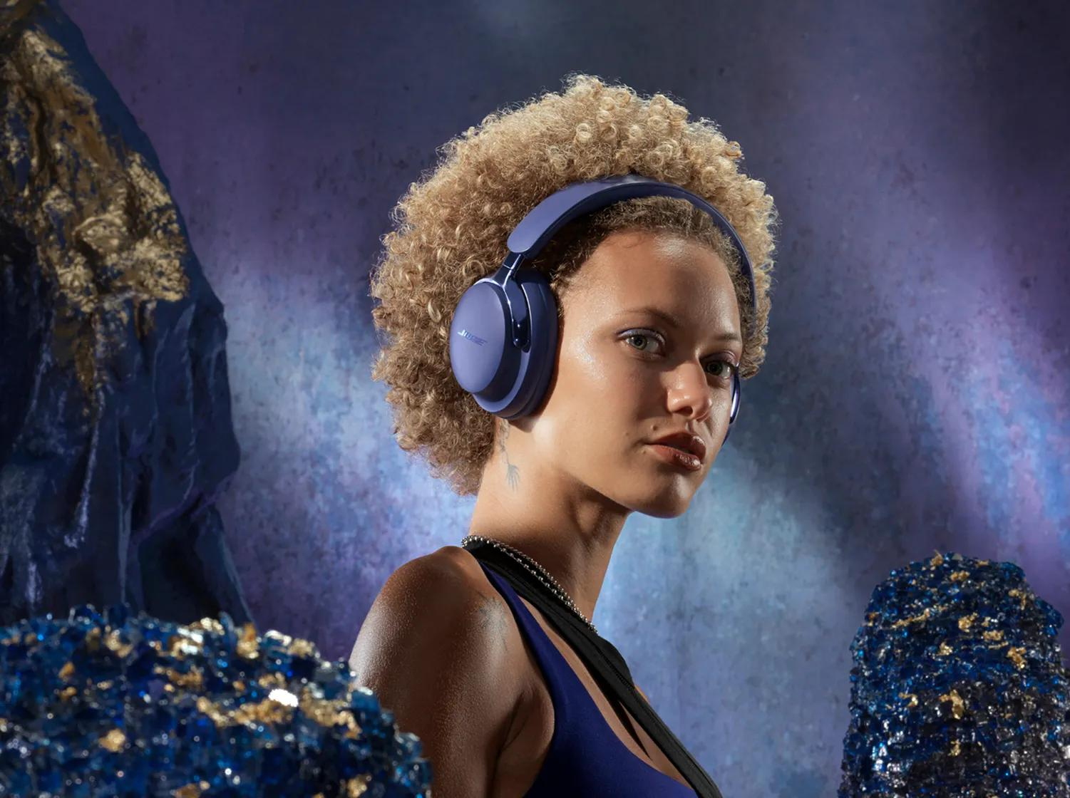 Kobieta z kręconymi włosami nosząca słuchawki. Ma na sobie niebieski top i naszyjniki, na niebieskim i fioletowym tle.
