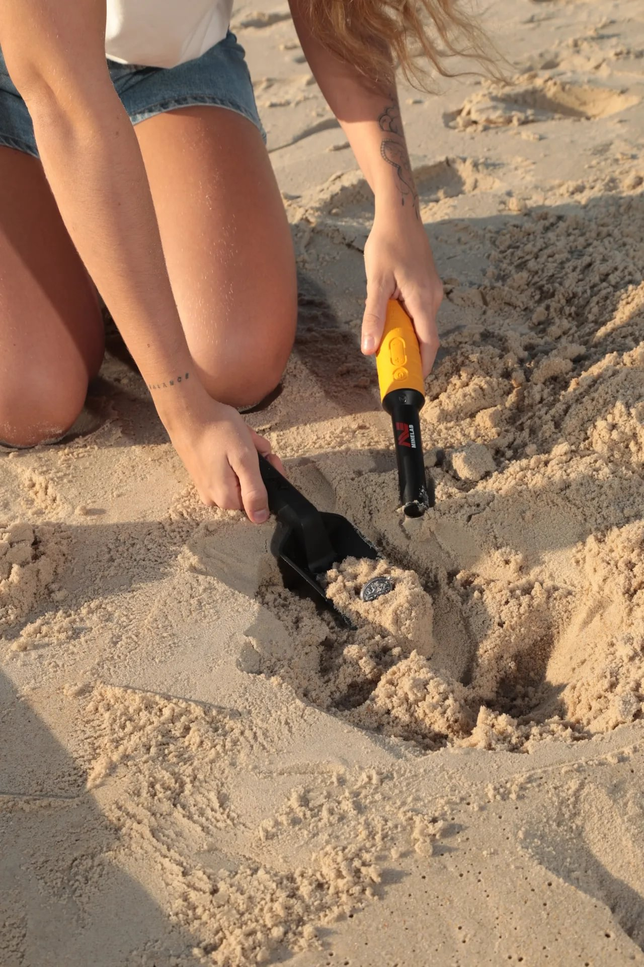 Person kniet am Strand und benutzt einen Metalldetektor und eine Schaufel, um etwas im Sand zu finden. Münze sichtbar.
