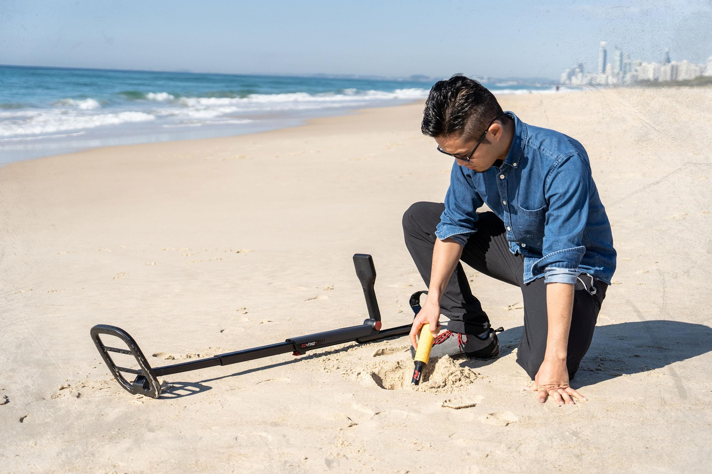 Ein Mann benutzt am Strand einen Metalldetektor. Meer und Gebäude sind im Hintergrund.