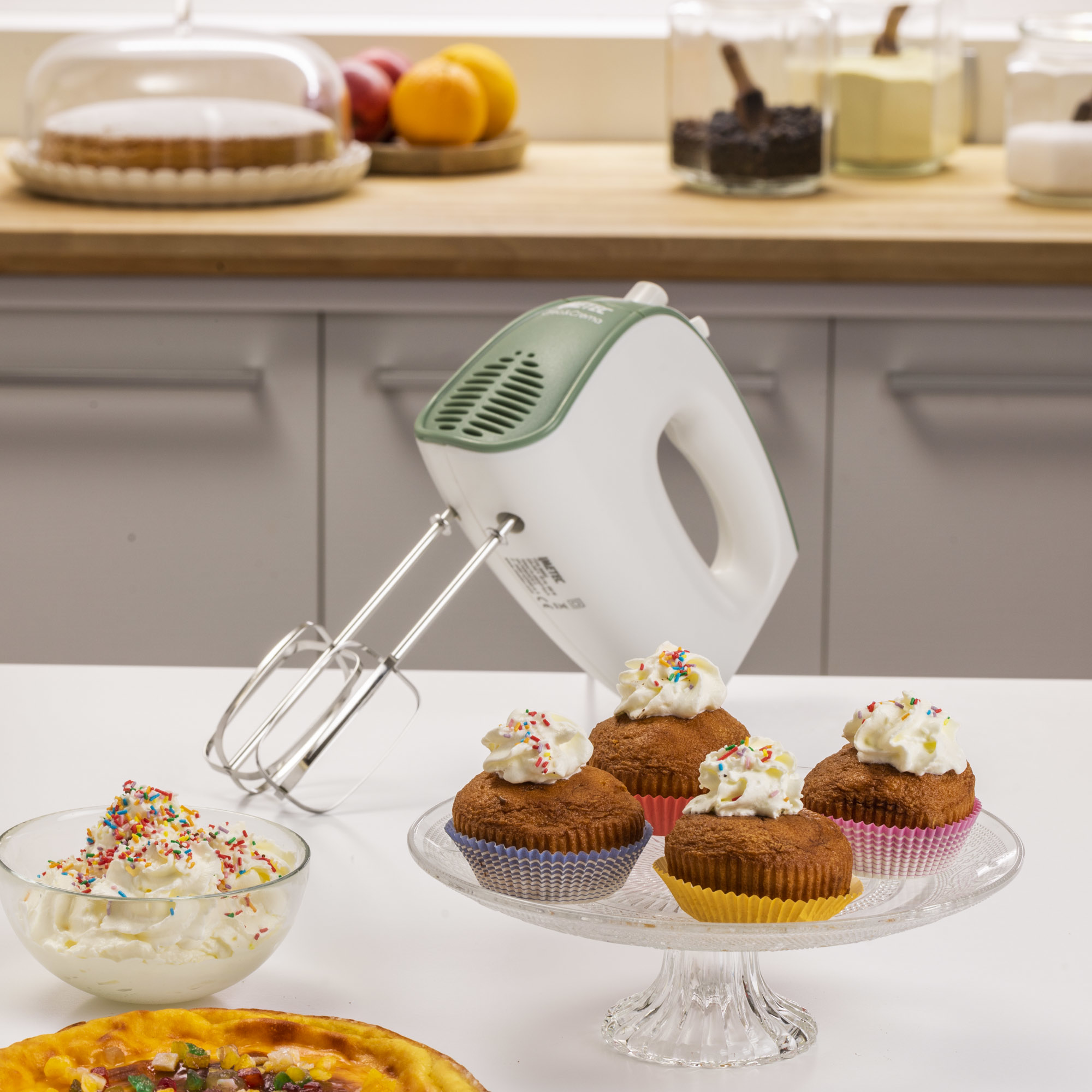 Escena de cocina con cupcakes, crema batida, batidora y pastel. Los cupcakes están en un soporte de pastel de vidrio.