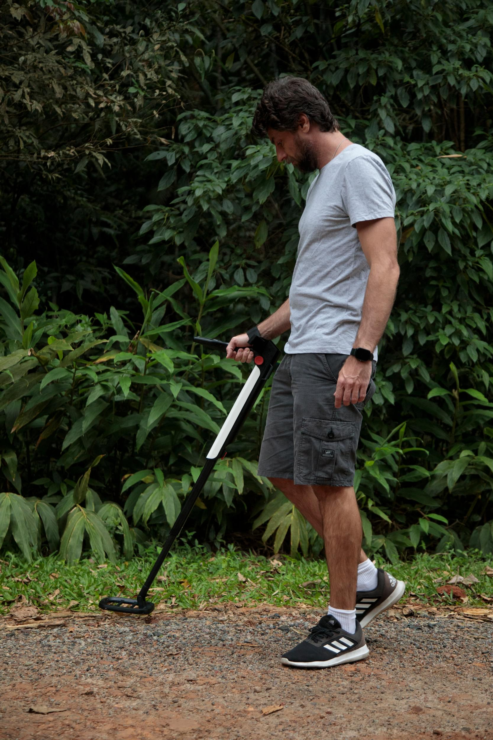 Uomo con metal detector cammina su un sentiero. Fogliame verde sullo sfondo. Indossa maglietta grigia, pantaloncini e scarpe da ginnastica.