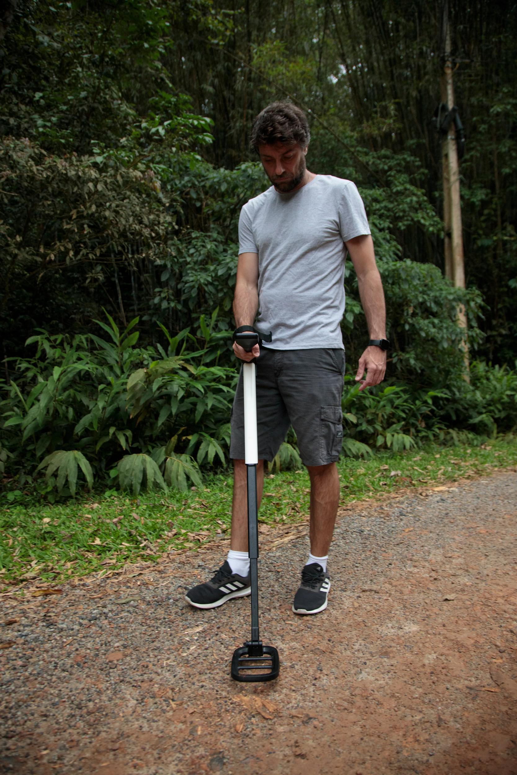 Uomo con metal detector nella foresta. Indossa una maglietta grigia, pantaloncini e scarpe nere.