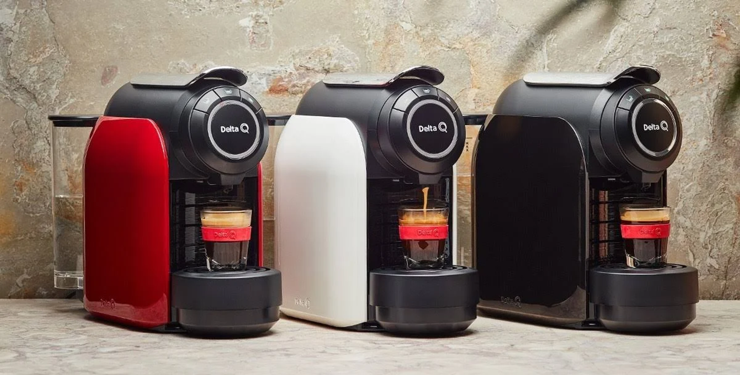 Tres máquinas de café Delta Q, rojas, blancas y negras. El café se vierte en un vaso. El fondo es una pared de piedra.