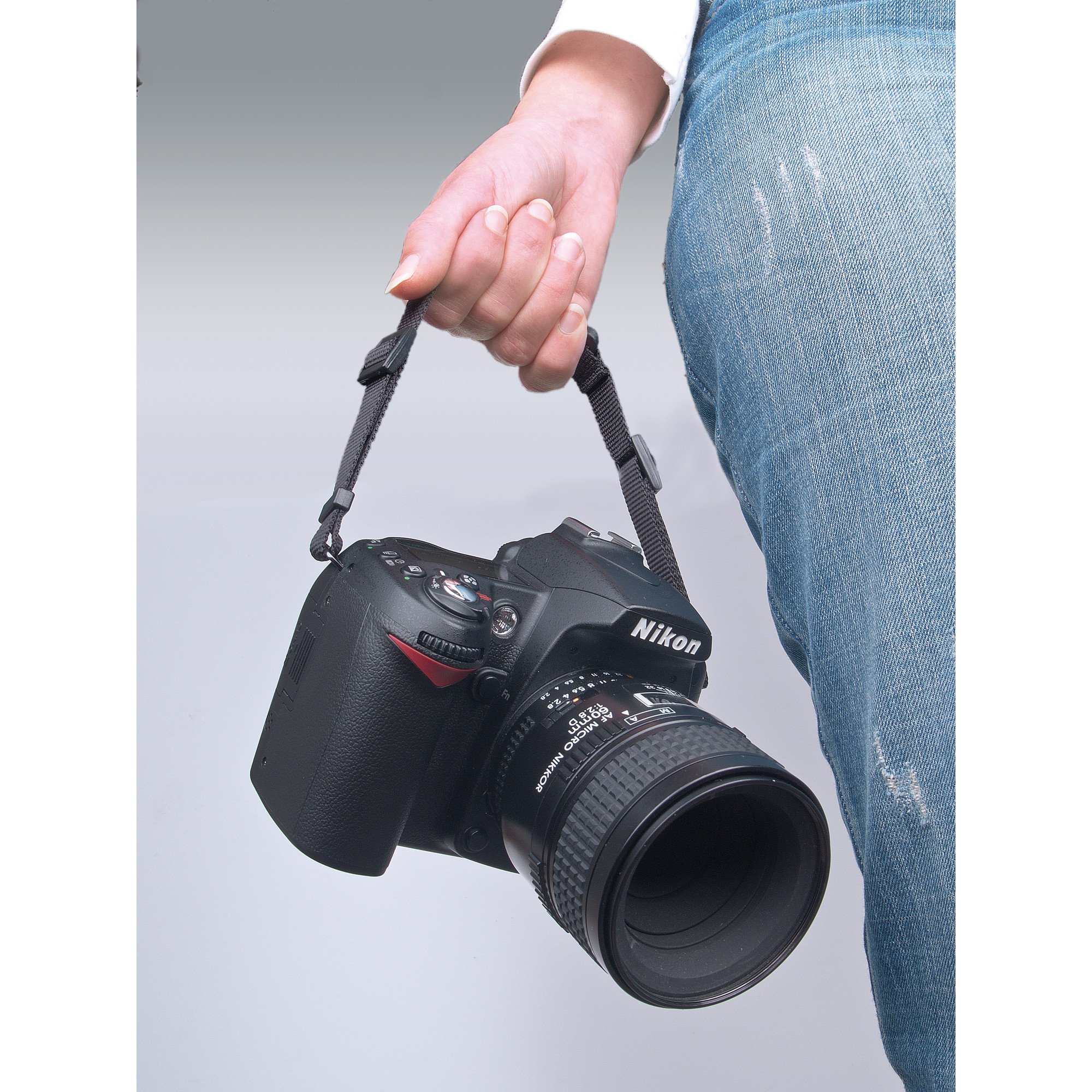 Eine Hand hält eine schwarze Nikon DSLR-Kamera an ihrem Riemen. Blaue Jeans und ein weißer Ärmel sind teilweise sichtbar.