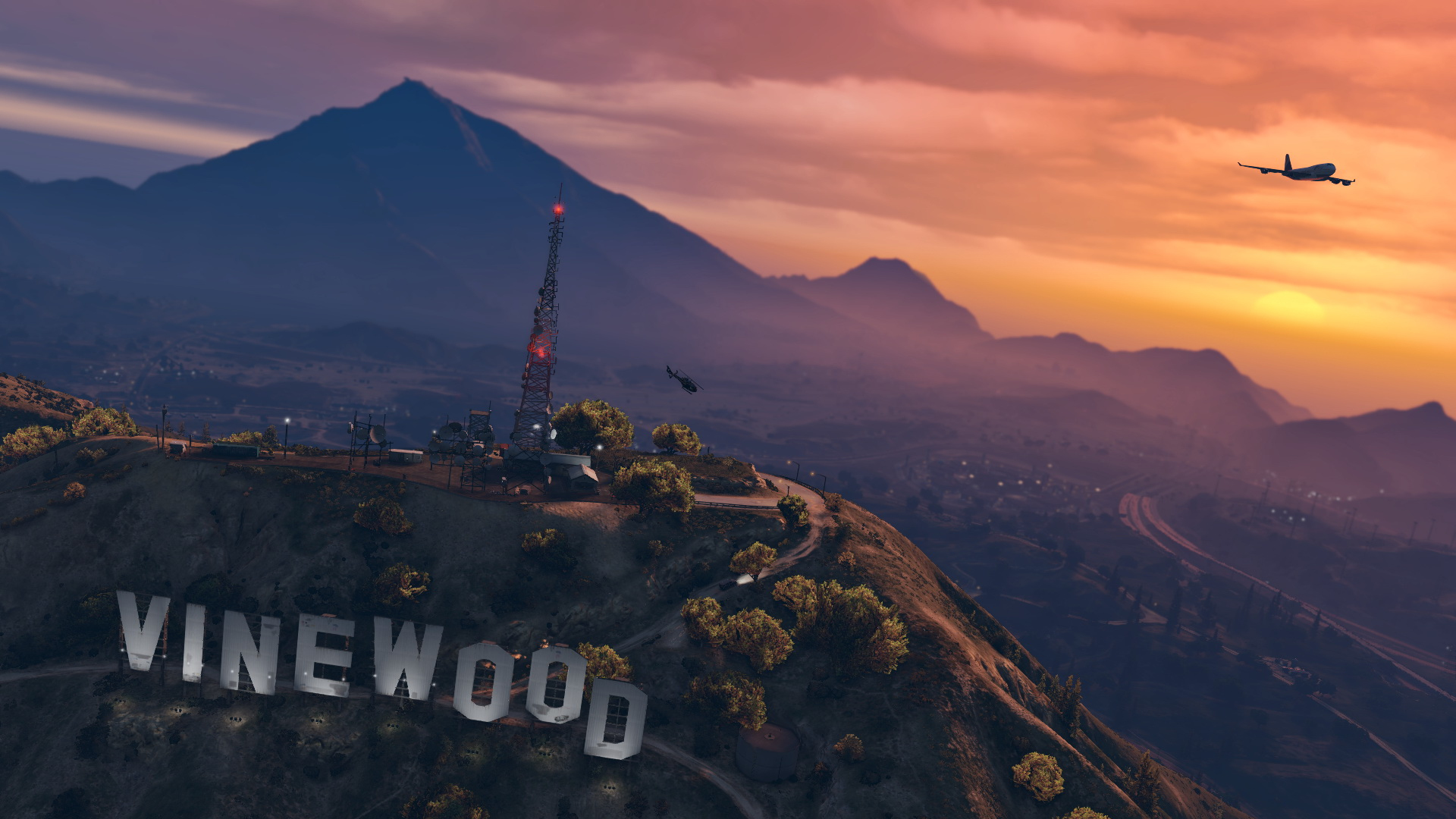 Cartello VineWood su una collina, tramonto con montagne, un aereo e un elicottero nel cielo, luci della città sotto.