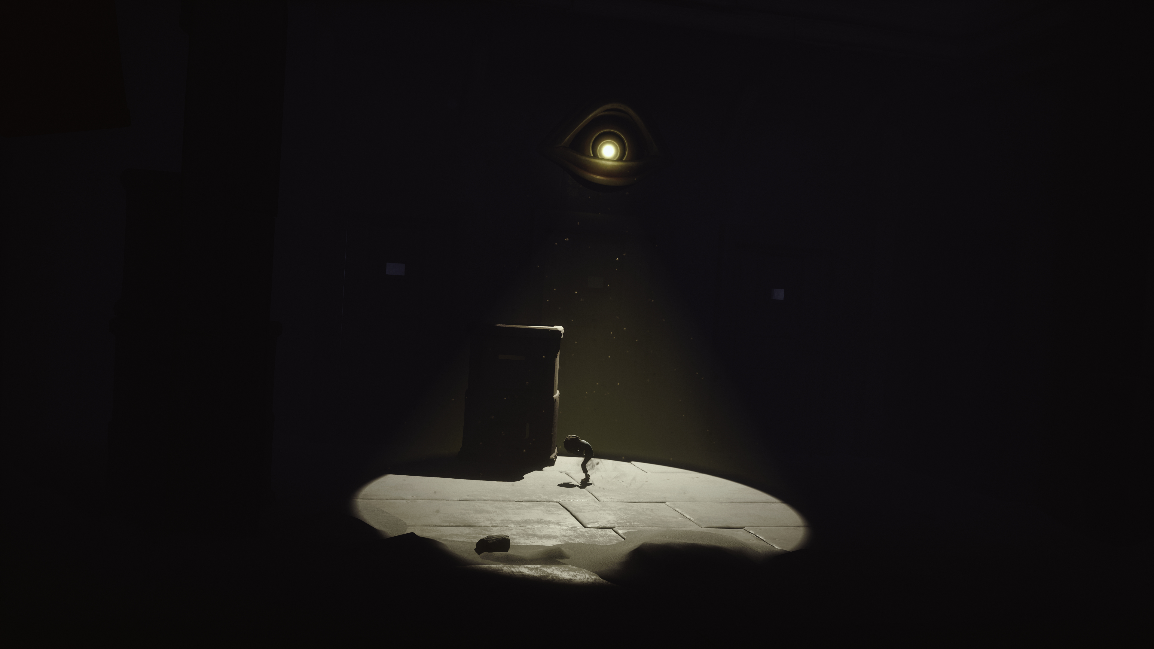 Una stanza buia con un riflettore. Una figura si inginocchia davanti a una scatola. Sopra, una forma di occhio luminoso illumina la scena.