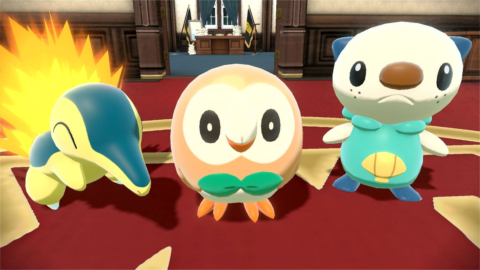 Tre personaggi Pokémon: Cyndaquil con fiamme, Rowlet e Oshawott, in una stanza con una scrivania e bandiere.