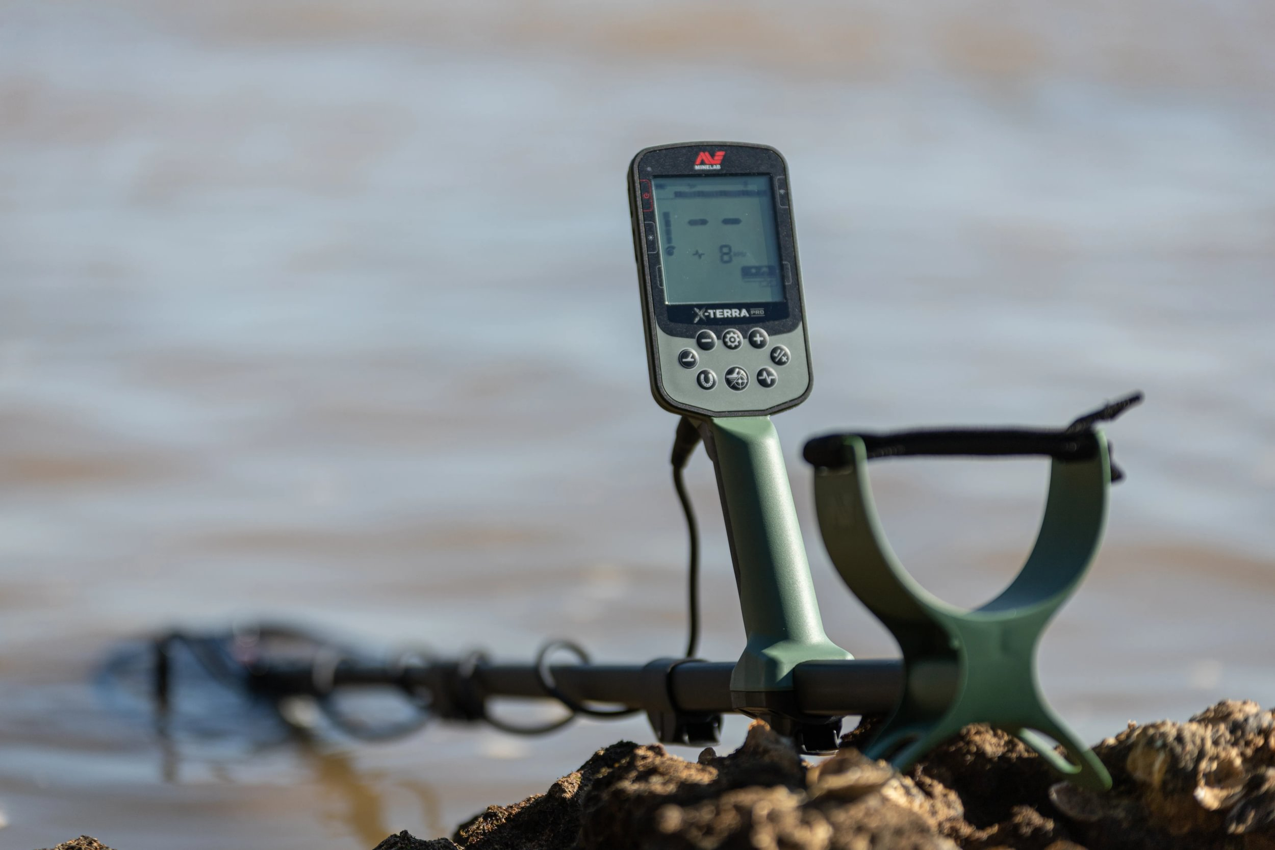 Ein Metalldetektor liegt auf einem Felsen in der Nähe von Wasser. Der Bildschirm zeigt Messwerte an. Der Detektor ist grün und schwarz.
