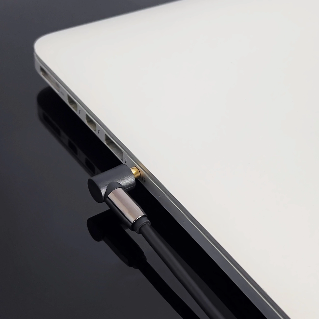 Ein Laptop mit eingestecktem Kabel. Das Kabel hat einen metallischen und schwarzen Stecker. Der Hintergrund ist eine glänzende schwarze Oberfläche.