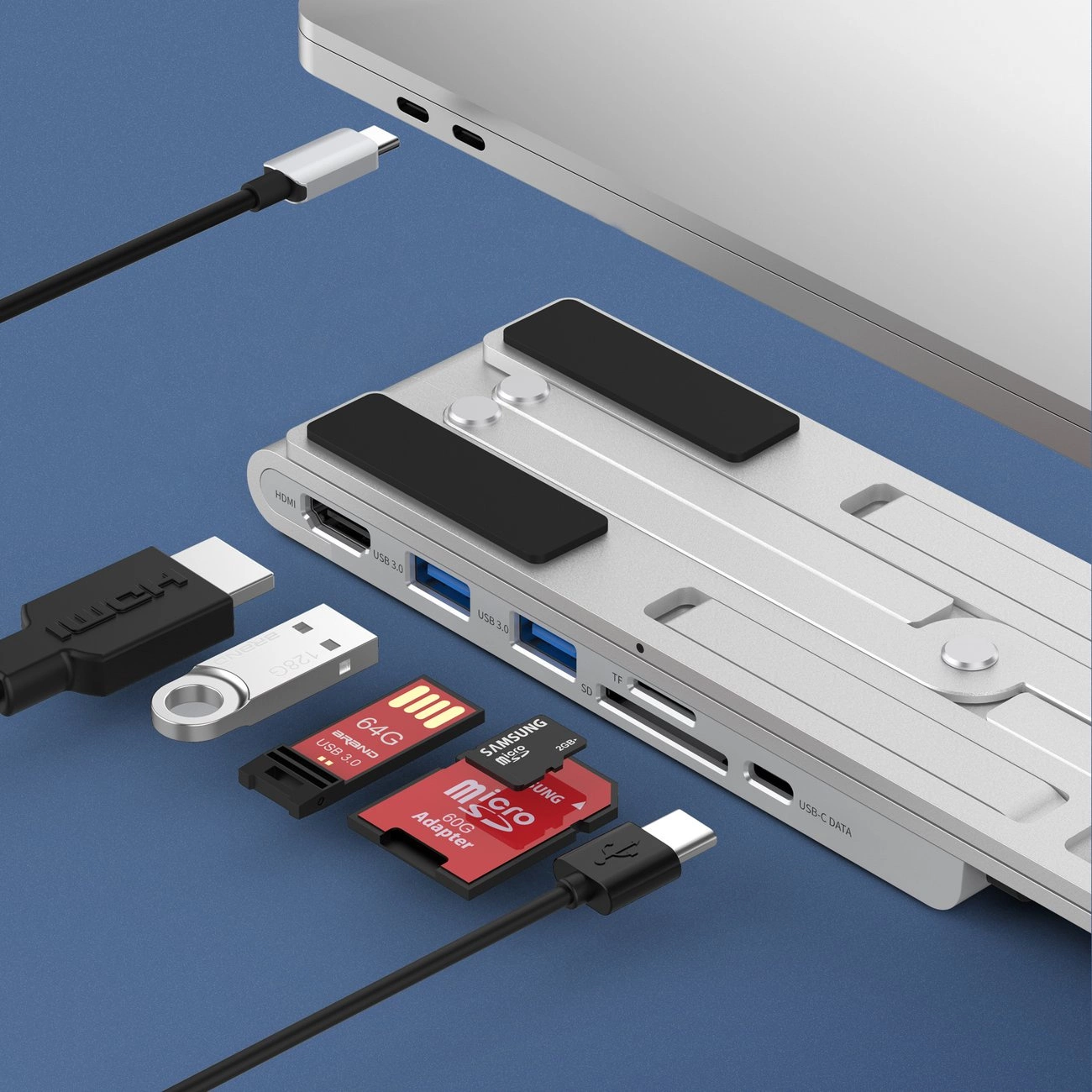 Ein Laptop mit einer silbernen Dockingstation, USB-Laufwerken und SD-Karten liegt auf einer blauen Oberfläche. Kabel verbinden sie.