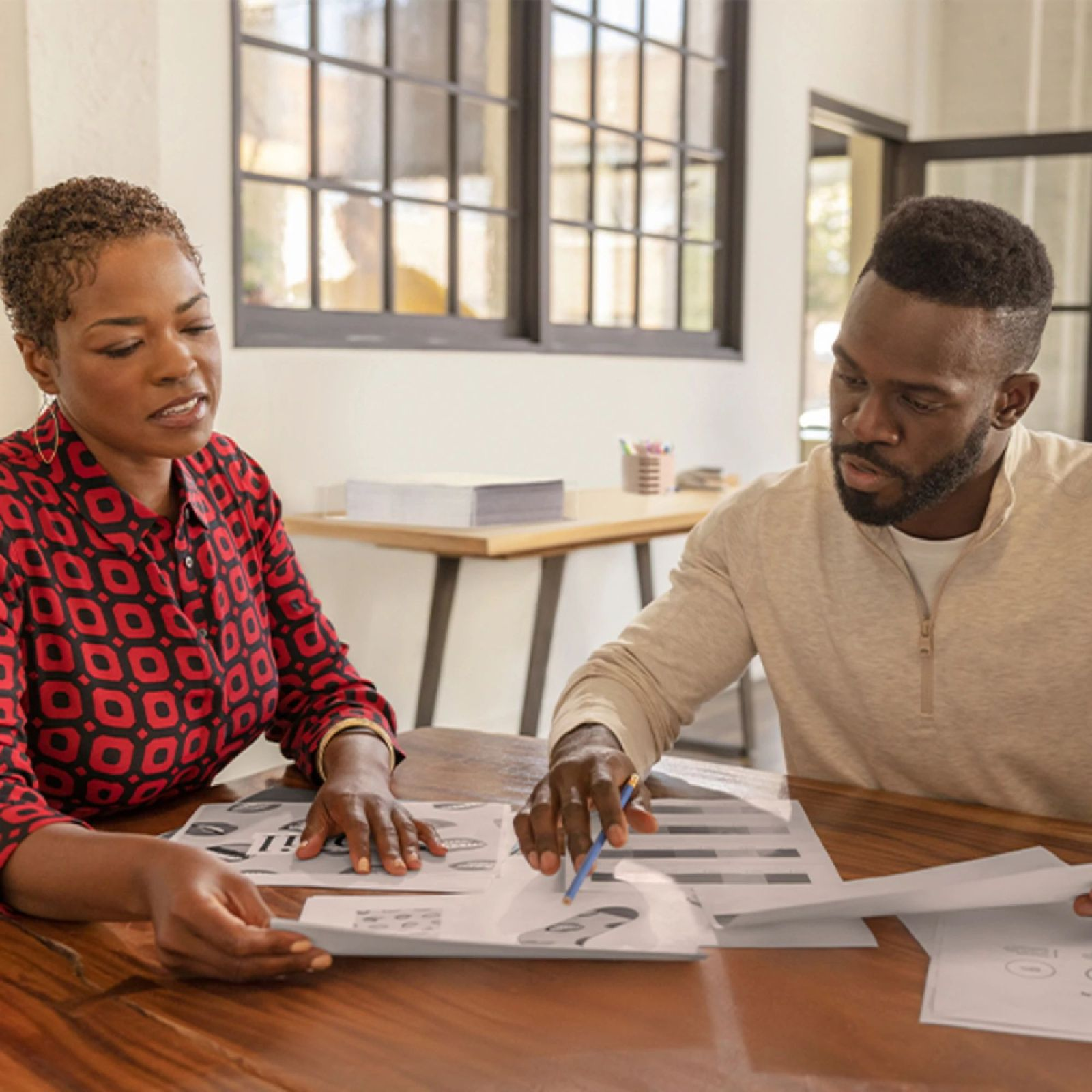Dwie osoby, mężczyzna i kobieta, siedzą przy stole i oglądają papiery. Kobieta ma na sobie czerwoną koszulę we wzory.