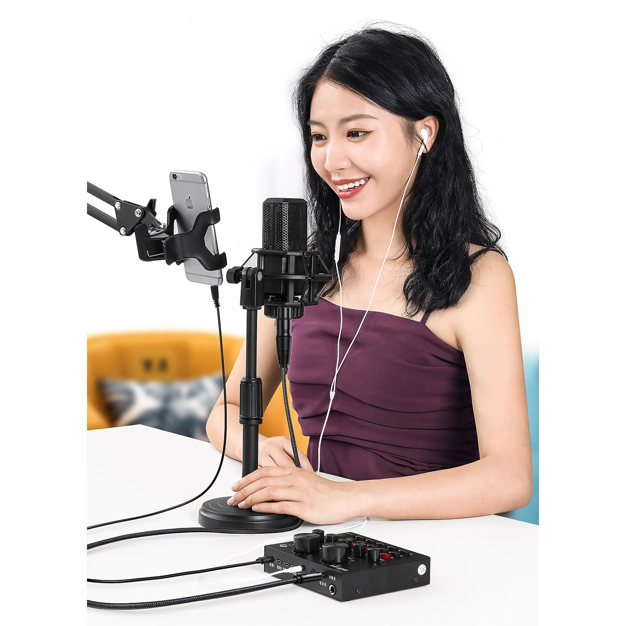Kobieta z mikrofonem i telefonem nagrywa. Uśmiecha się, ma na sobie fioletowy top i słuchawki. Czarny mikser audio na stole.