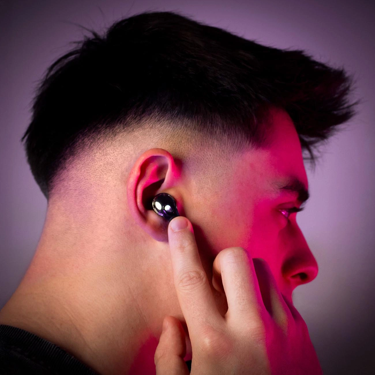 Un uomo con i capelli neri che indossa un auricolare nero. Il suo dito tocca l'auricolare. Luce rosa e viola sul viso.