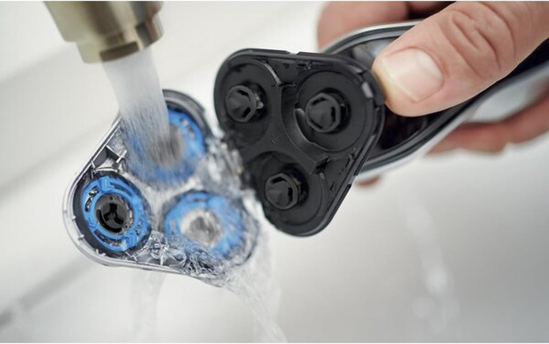 A close-up of an electric shaver being rinsed with water. The shaver head is black and blue, held by a hand.