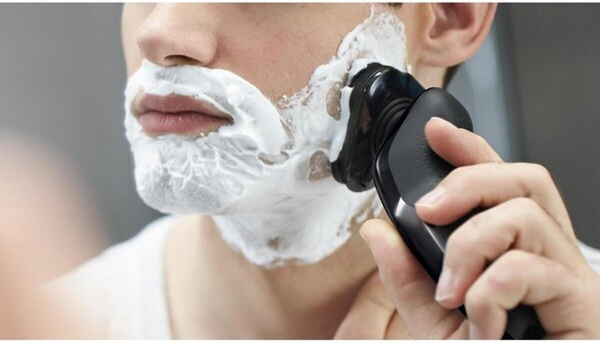 A man shaves his face with an electric razor. His face is covered in white shaving cream.